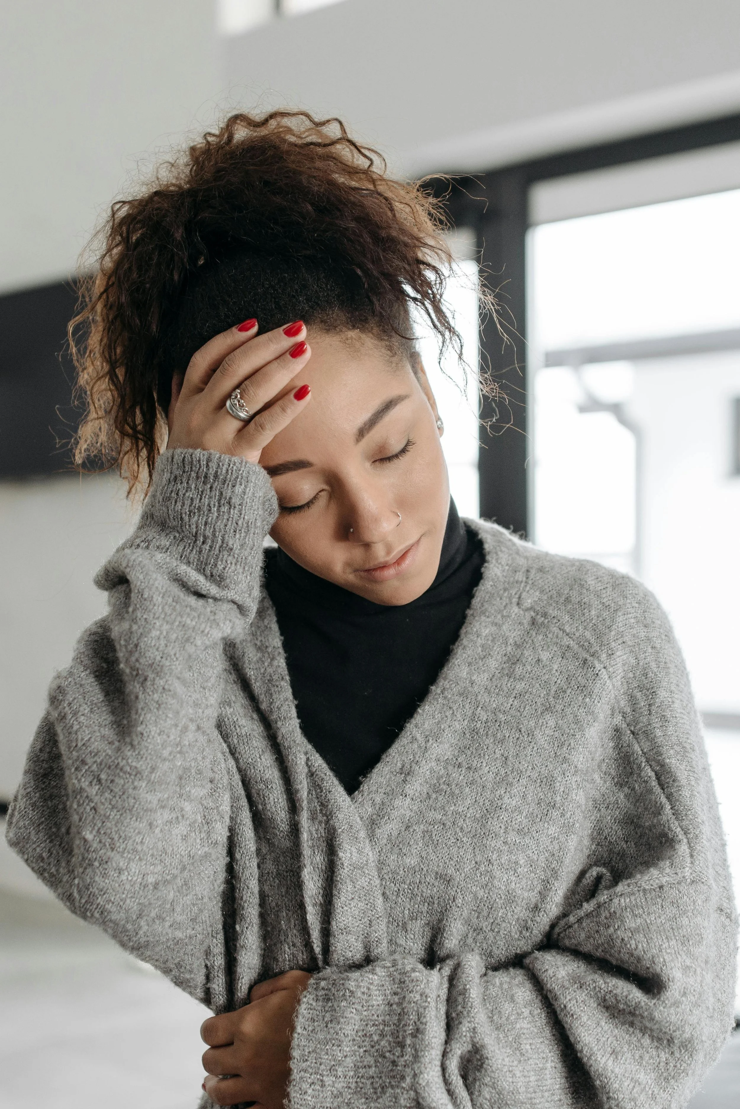 A woman with curly brown hair and light skin, wearing a grey sweater and black top, with her eyes closed and hand on her forehead, appearing to be in pain, standing in front of large windows with natural light.