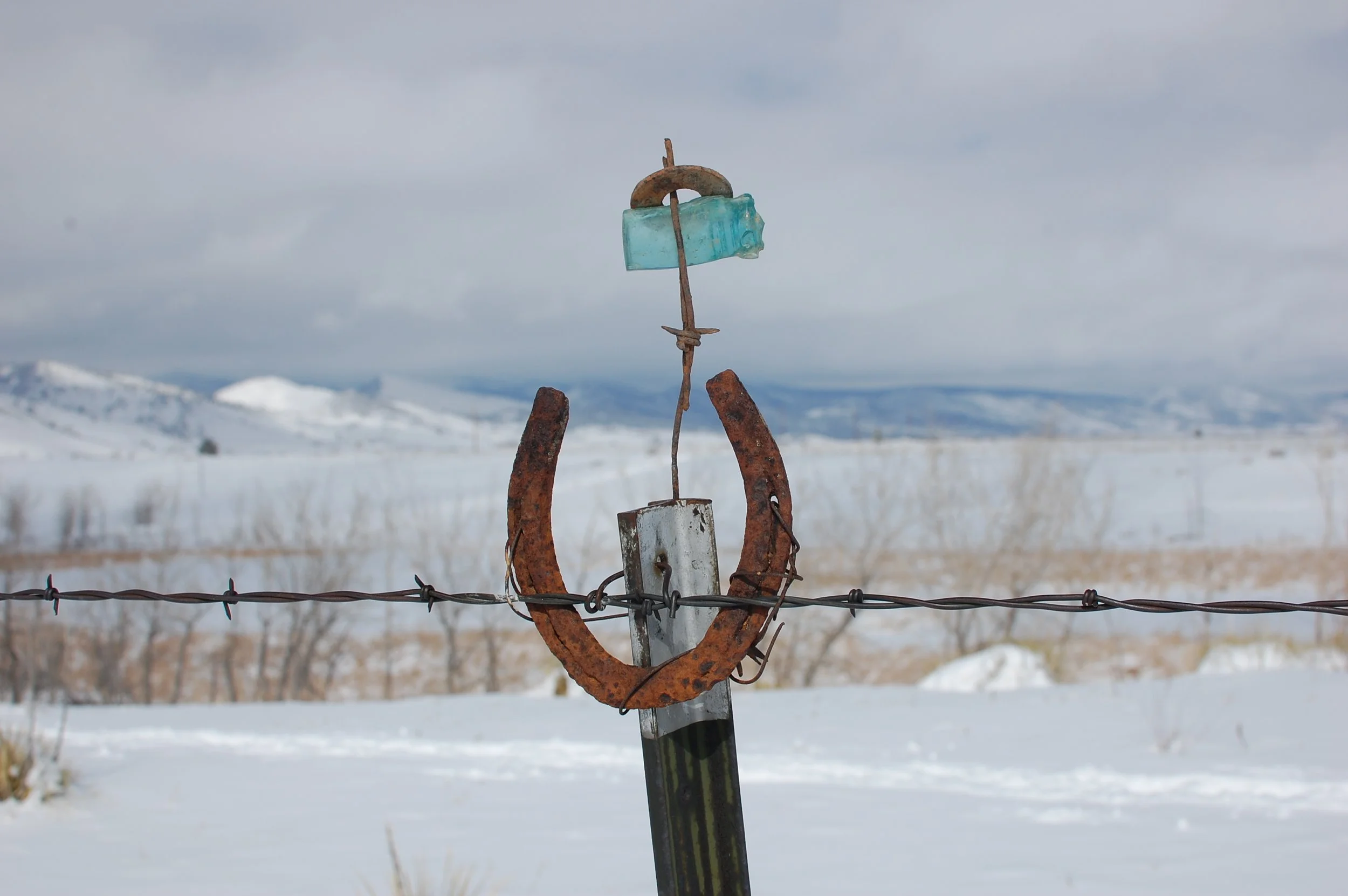 A horseshoe decorated with turquoise blue glass on a barbed-wire fence against a snowy landscape with mountains and cloudy sky in the background.