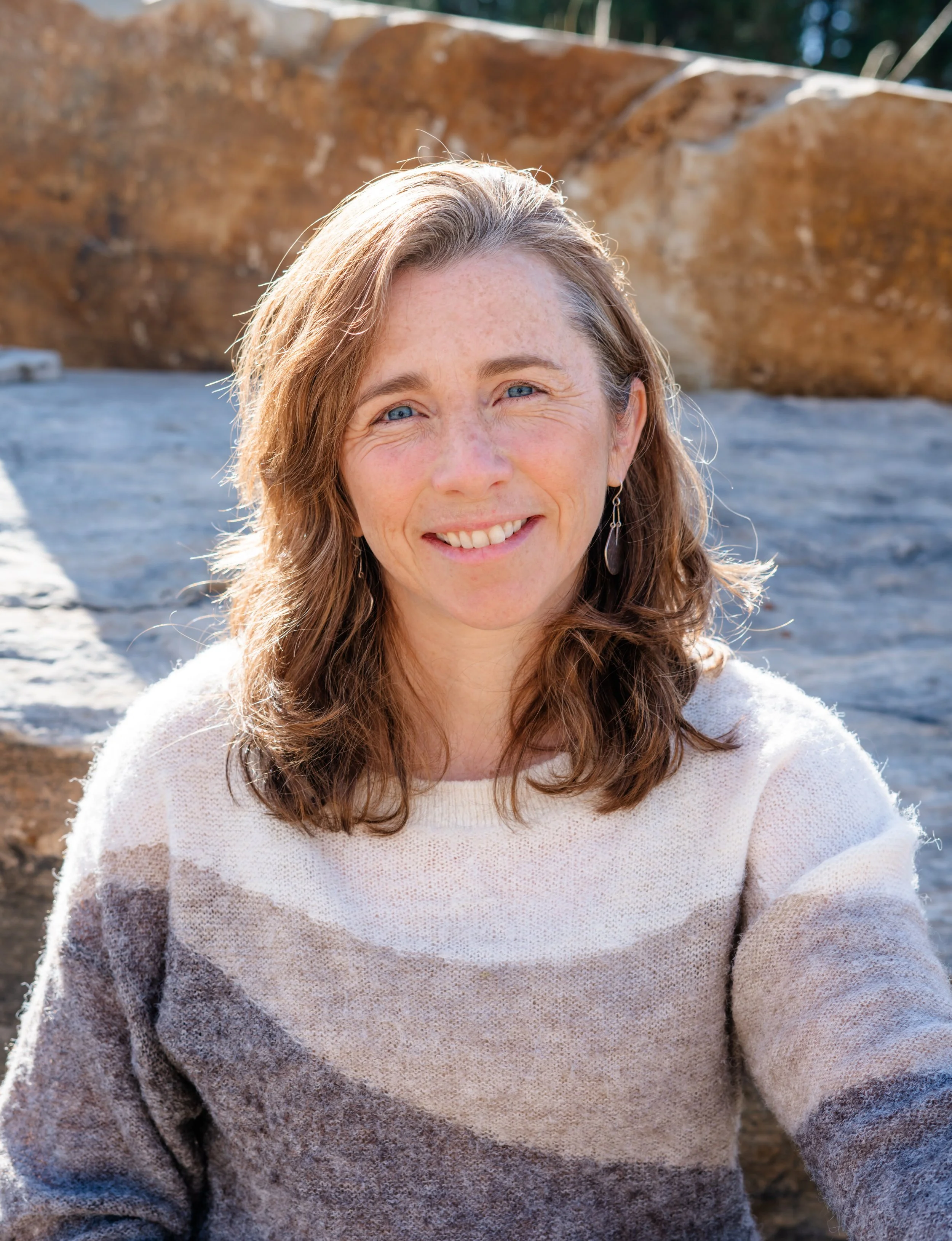 Close-up of a smiling woman with wavy brown hair and blue eyes, wearing a white sweater, outdoors with rocks and trees in the background.