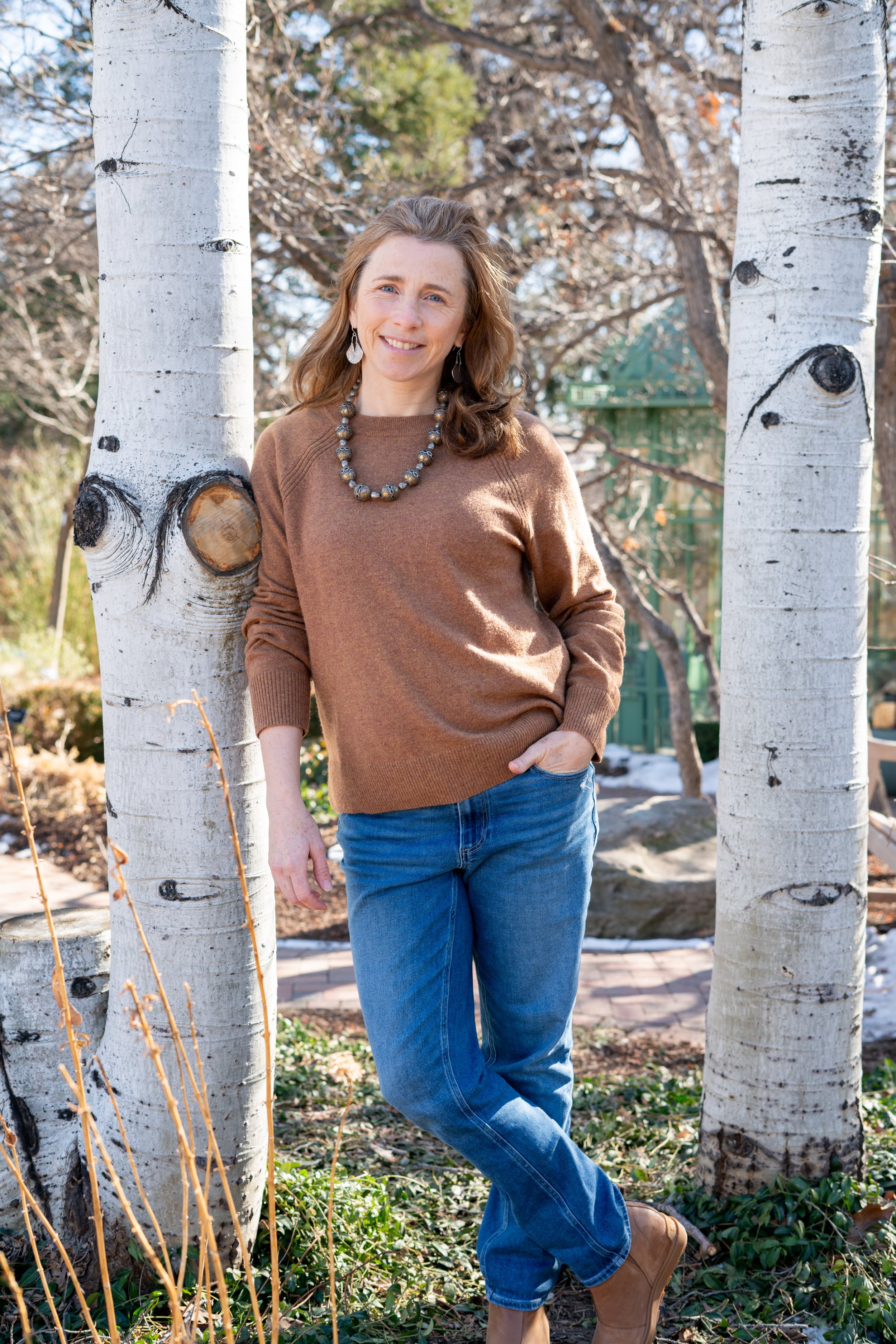 A woman standing outdoors between two white birch trees, smiling, wearing a brown sweater, blue jeans, and tan boots, with a necklace and earrings, on a sunny day with aspen trees in the background.