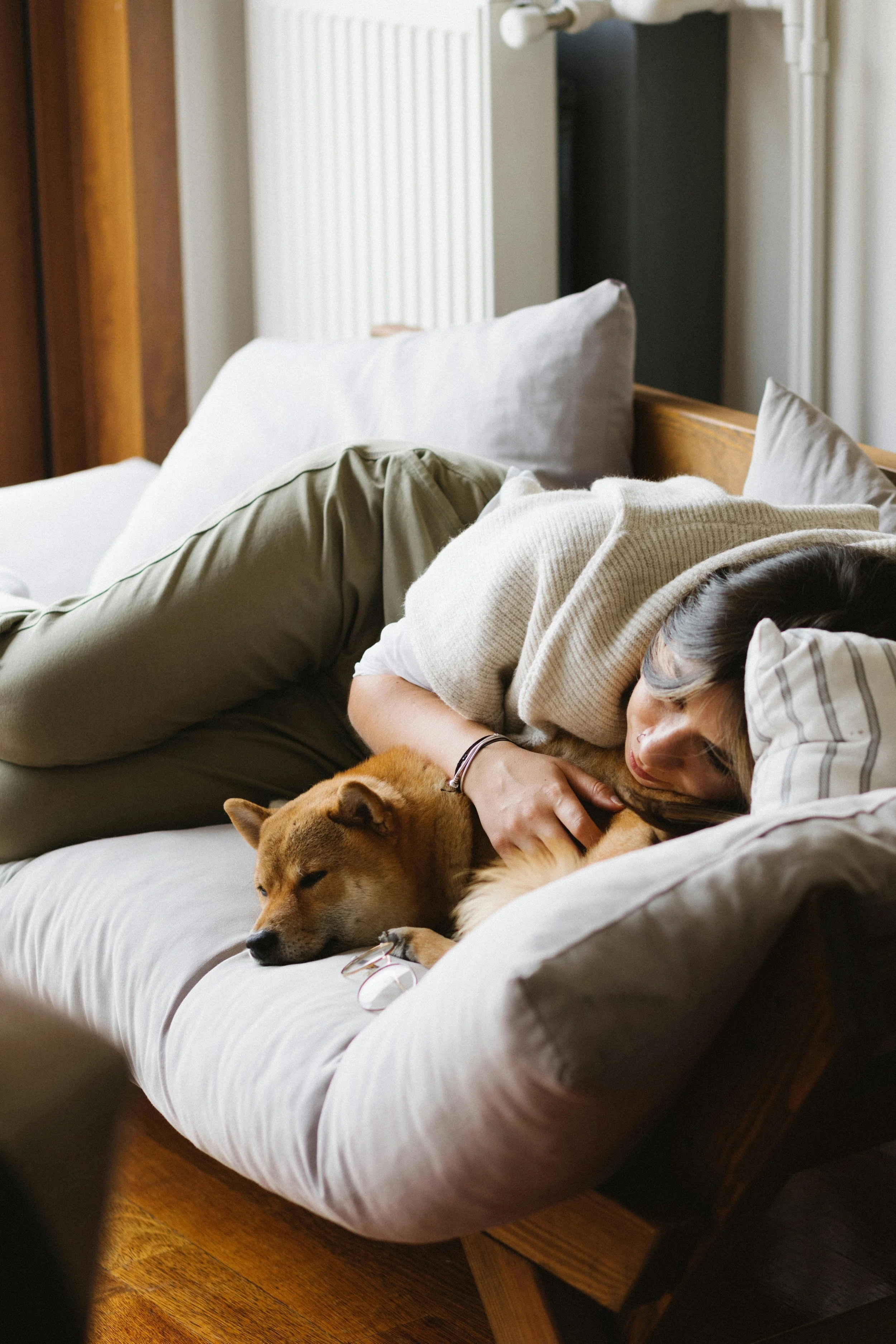 A woman lying on her side on a bed with two large pillows, cuddling a golden retriever dog. The dog is lying on the bed with its head resting on the mattress. The woman is wearing a beige sweater and green pants, with her face partially obscured as she rests her head on a striped pillow. The room has wooden floors and a white radiator in the background.