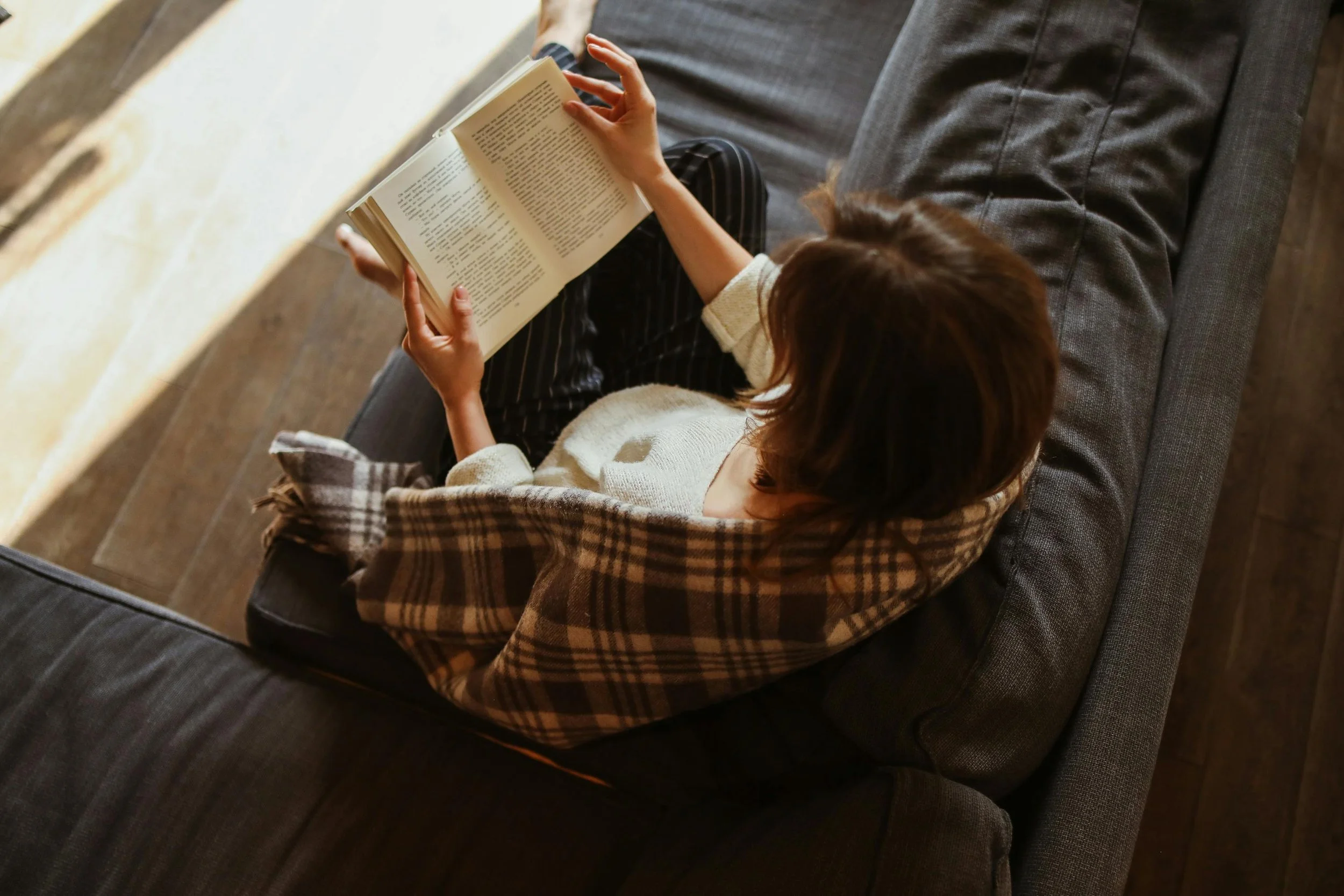 A person with brown hair sitting on a dark gray couch, reading a book, dressed in a cream-colored sweater and striped pants, near a brick wall.