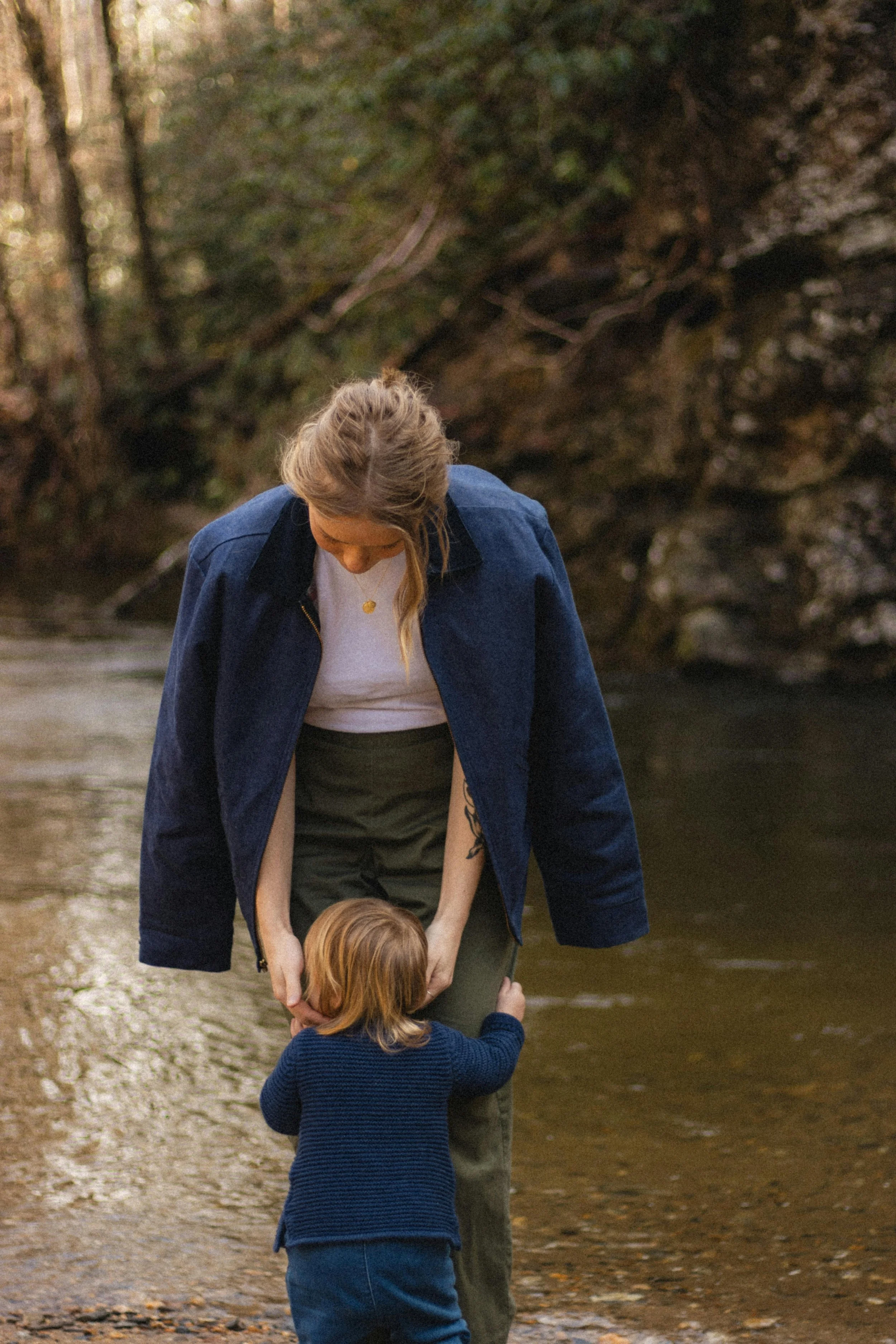 A woman with light brown hair, wearing a dark blue jacket over a white shirt, kneels by a young child with blonde hair, near a stream in a wooded area. The woman is looking down at the child, who is reaching out towards her. The scene is outdoors, surrounded by trees and natural scenery.