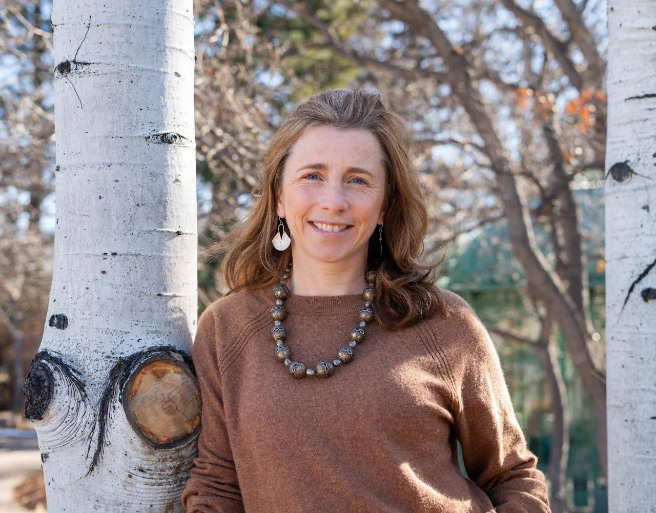A woman with shoulder-length brown hair, wearing earrings and a necklace, smiling outdoors next to a white aspen tree during daytime.