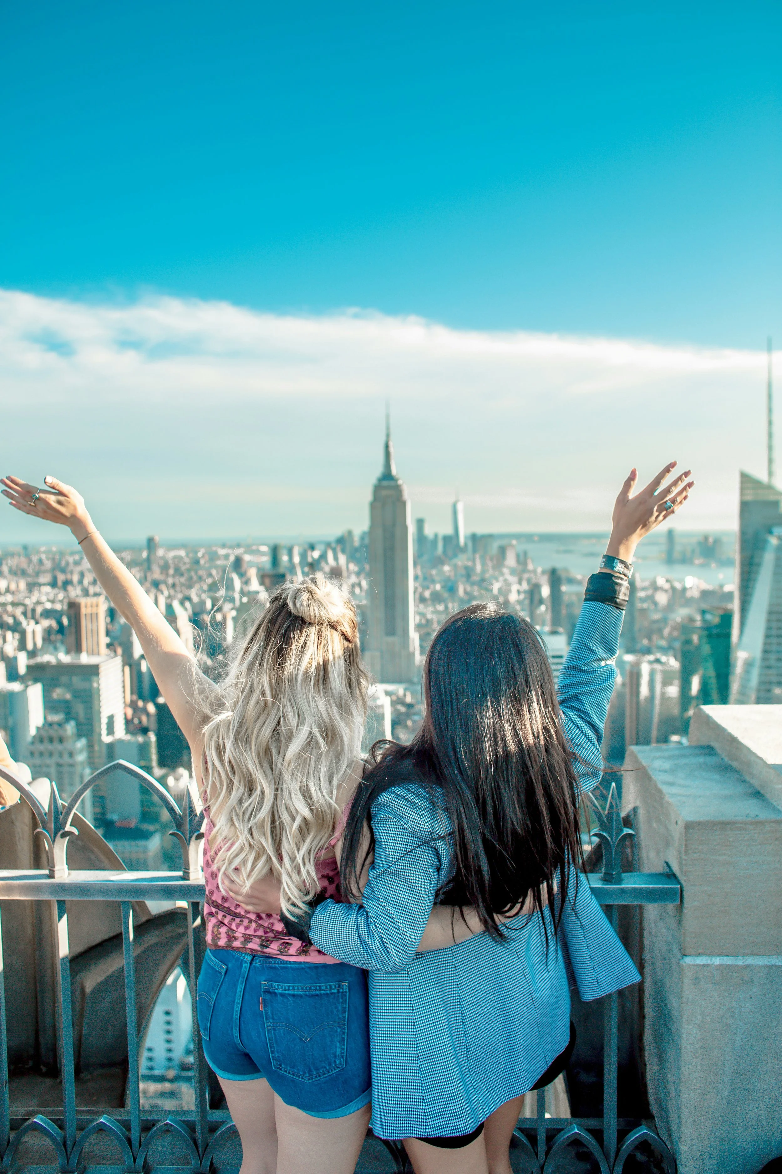 Two women with long hair, one with wavy blond hair and the other with straight black hair, embrace on a rooftop overlooking New York City with the Empire State Building in the background, during daytime.
