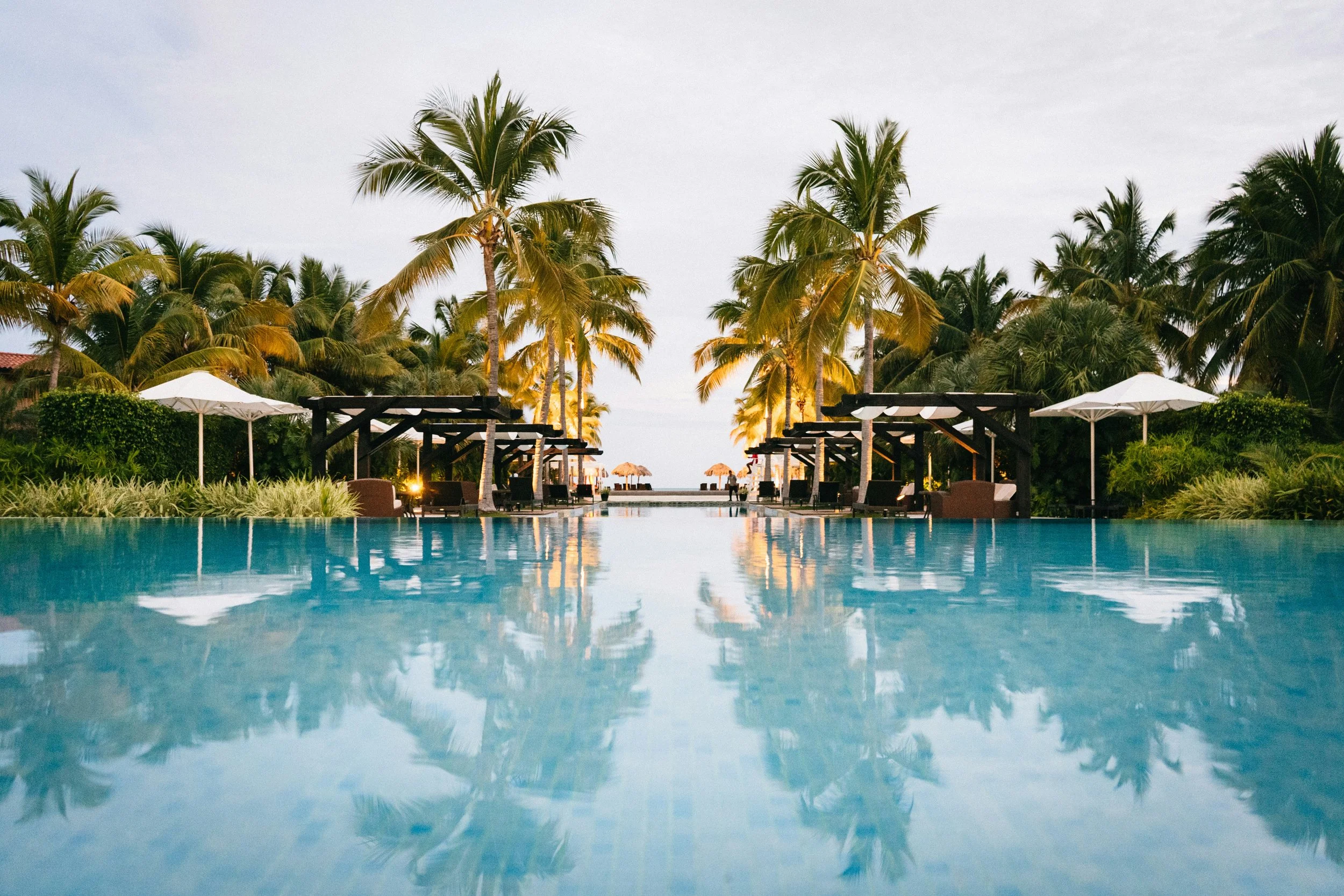 Poolside view with palm trees, white umbrellas, lounge chairs, and thatched umbrellas in the background, near the beach.
