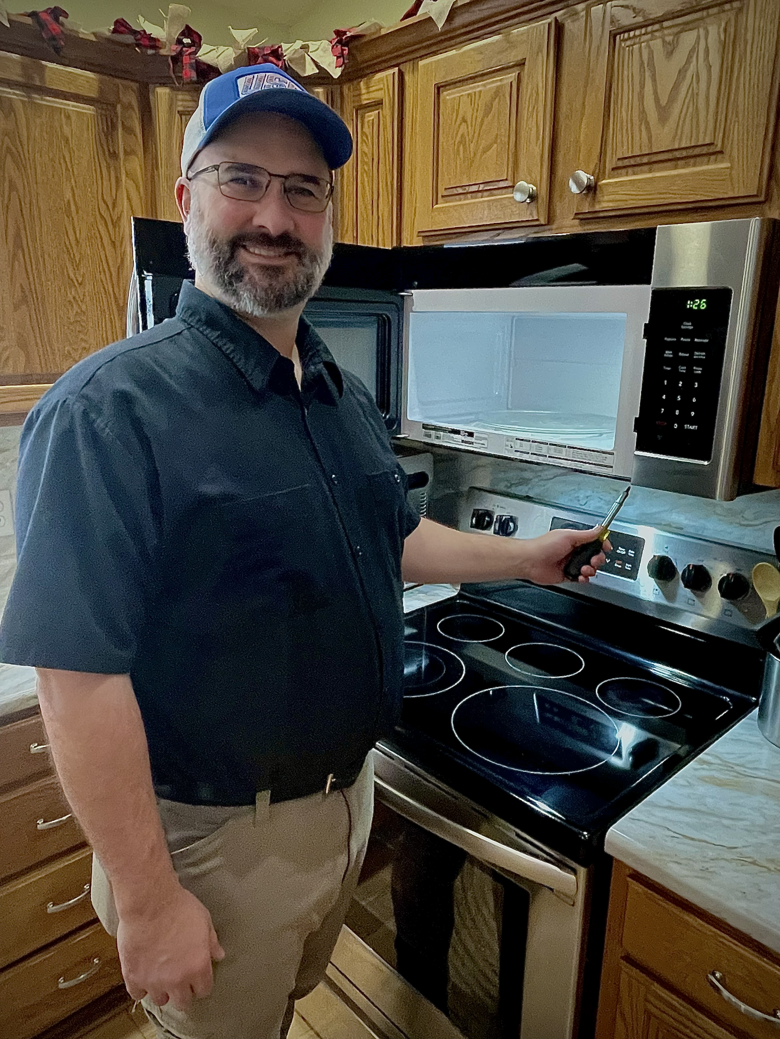 A man with glasses and a beard is standing in a kitchen, smiling at the camera. He is holding a screwdriver and pointing at the control panel of a microwave oven that is installed above a stove. The man is wearing a blue cap, a black short-sleeved shirt, and khaki pants. The kitchen has wooden cabinets and a marble countertop, with holiday decorations visible in the background.