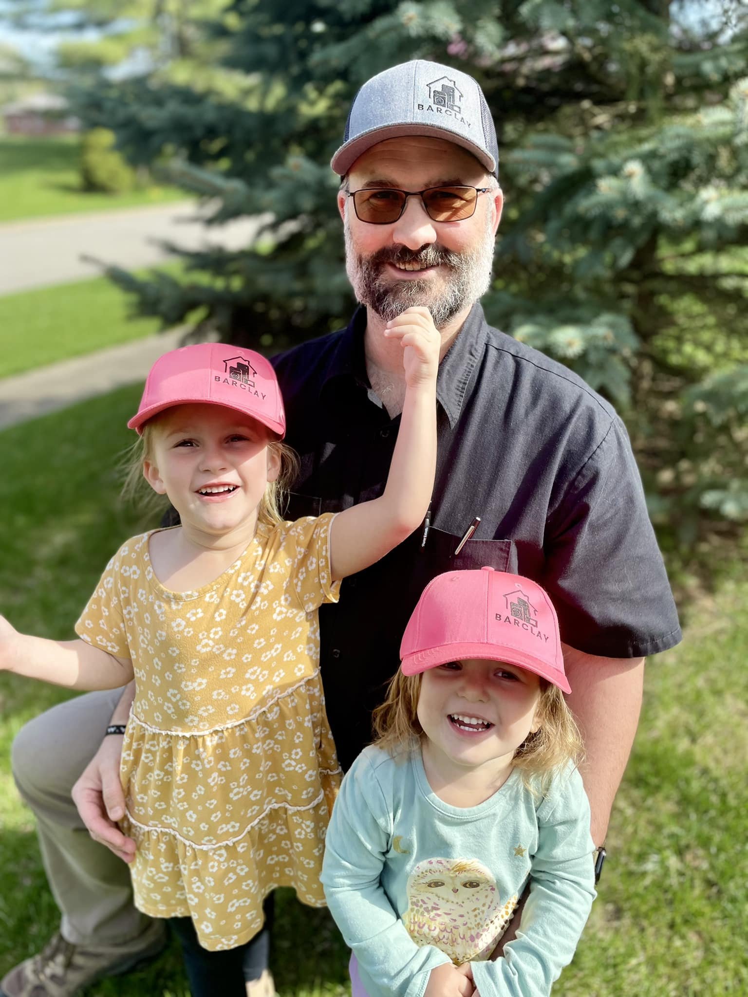 A man with a beard, glasses, and a gray baseball cap is outdoors with two young girls, all wearing pink caps. The man is wearing a black shirt and smiling. One girl has blond hair, is wearing a yellow dress with white flowers, and has her arm raised. The other girl has light brown hair, is wearing a mint green shirt with an owl graphic, and is smiling at the camera. They are standing on grass with trees in the background.