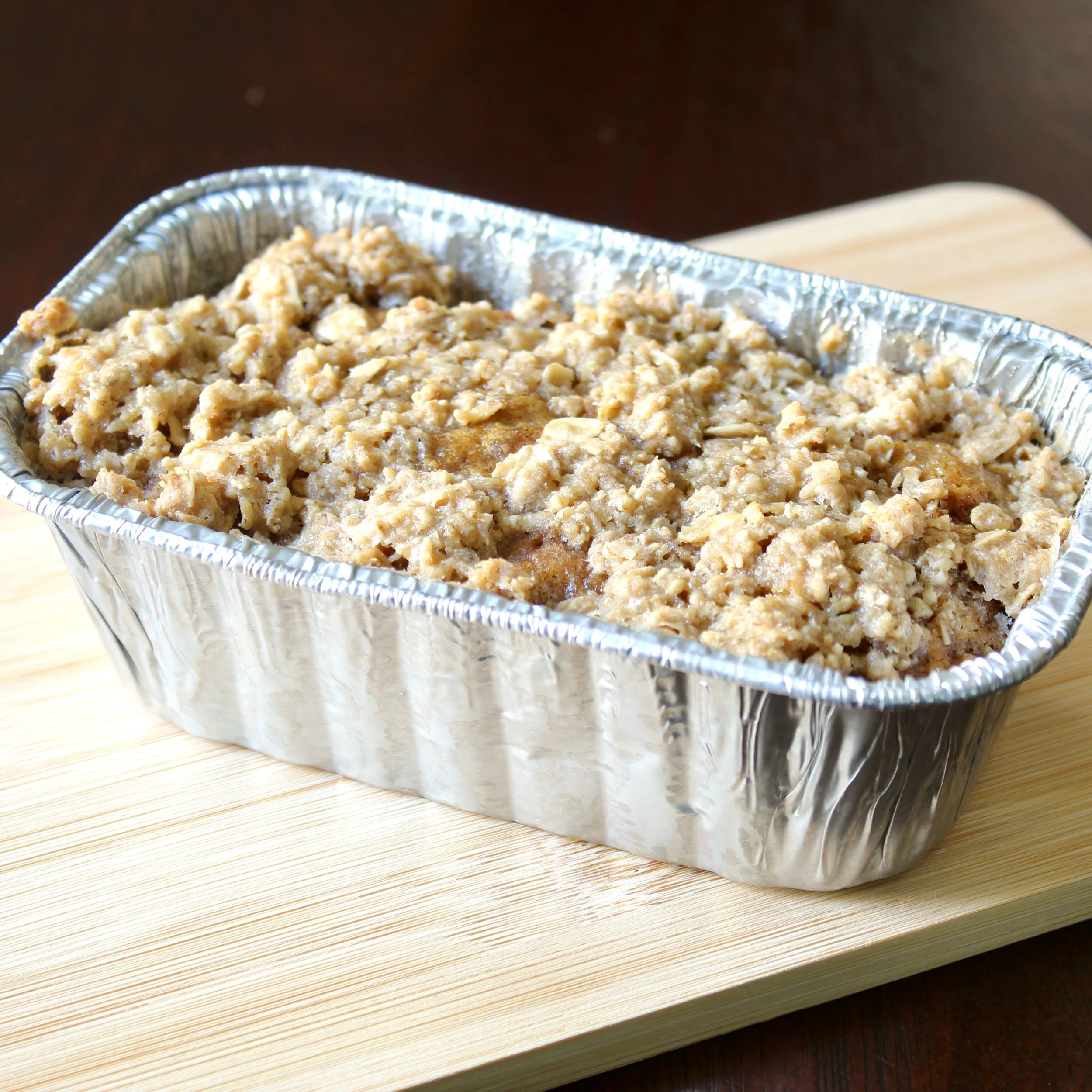 Streusel-topped pumpkin bread loaf baked in a foil tin on a light wooden board.