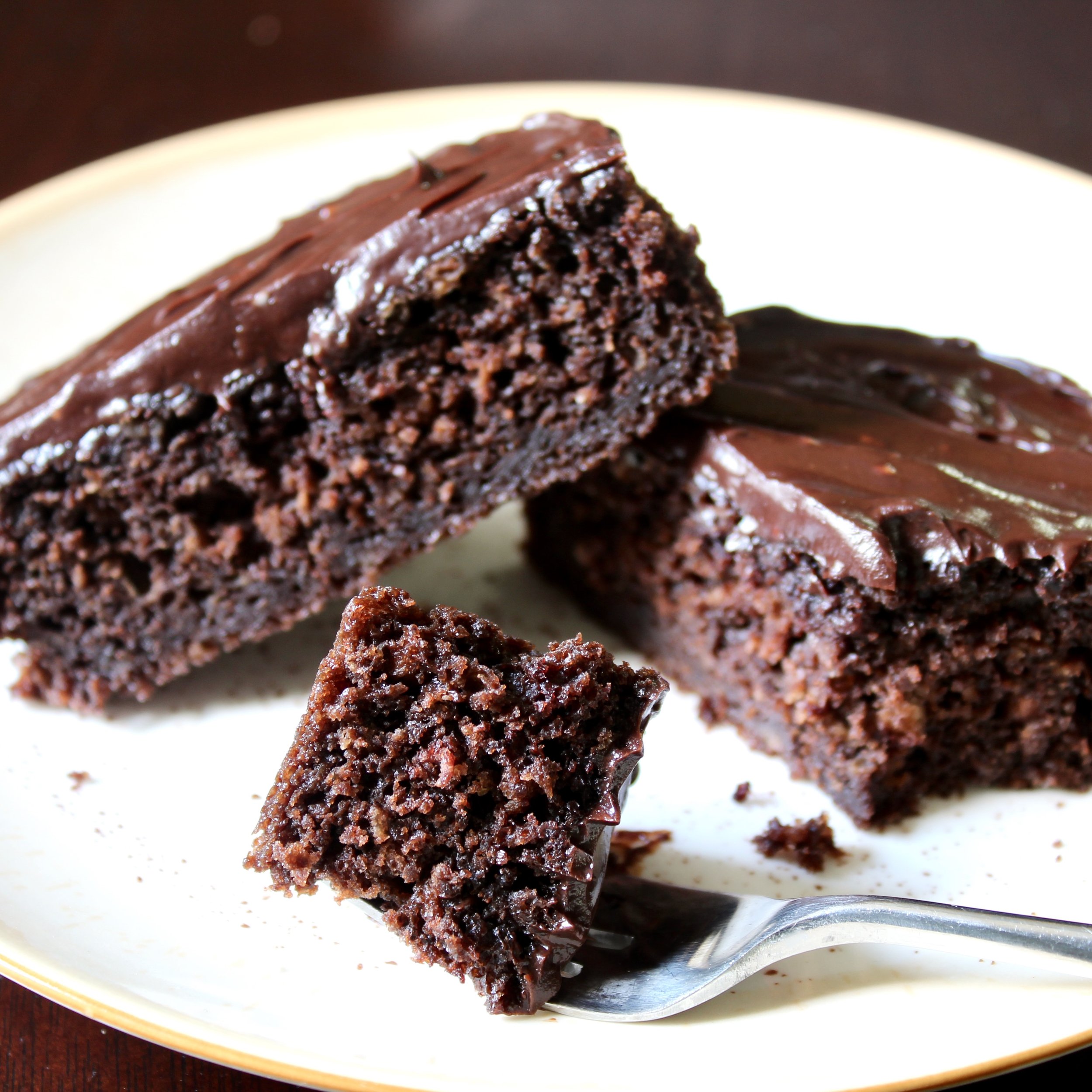 Slices of rich chocolate cake topped with dark chocolate ganache rest on a neutral plate, with a bite held forward on a silver fork.