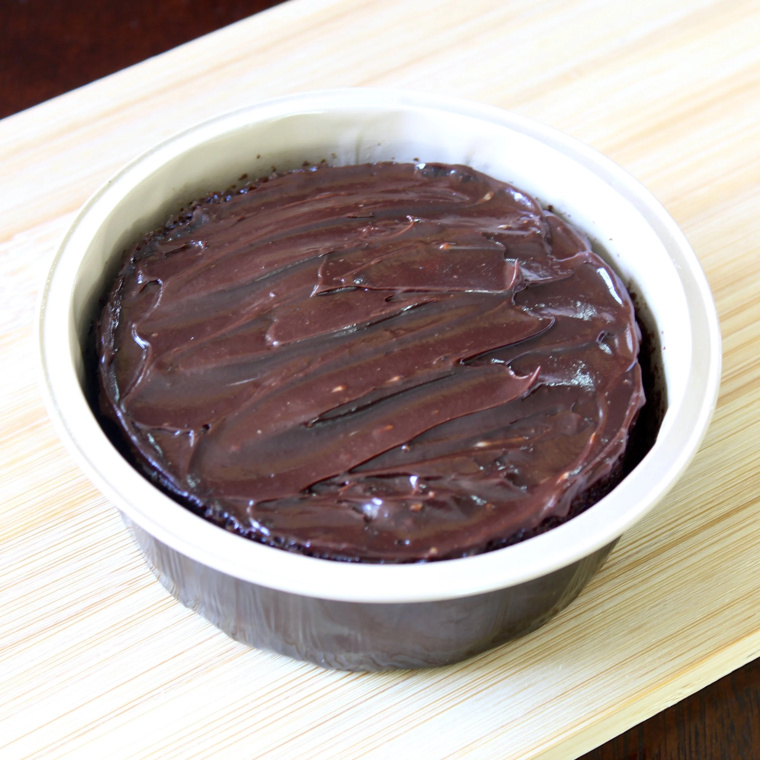Rich, frosted chocolate cake baked in an individual ramekin, styled on a minimal bamboo board.