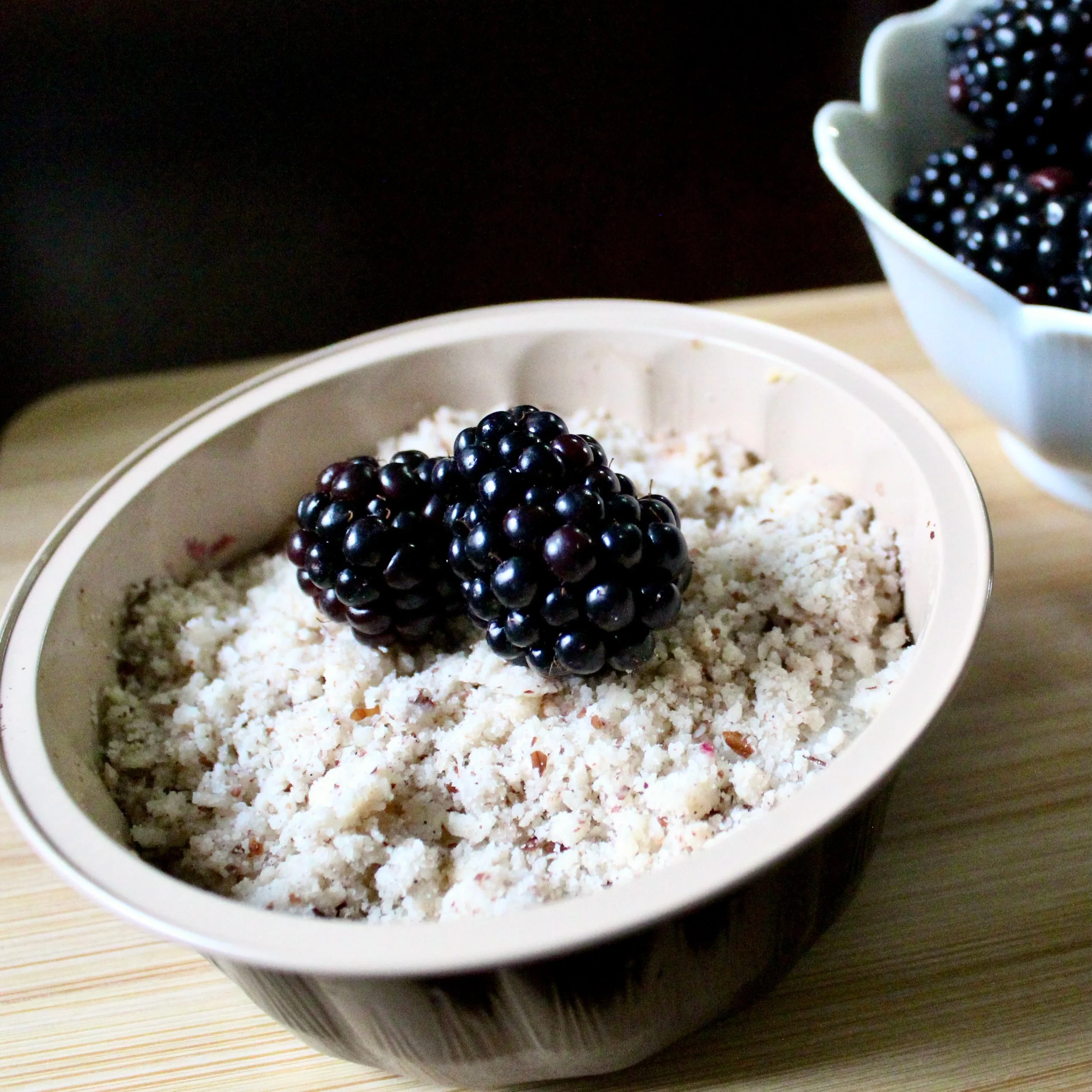 Two fresh blackberries top a pale crumble dessert inside a small, dark ceramic bowl.