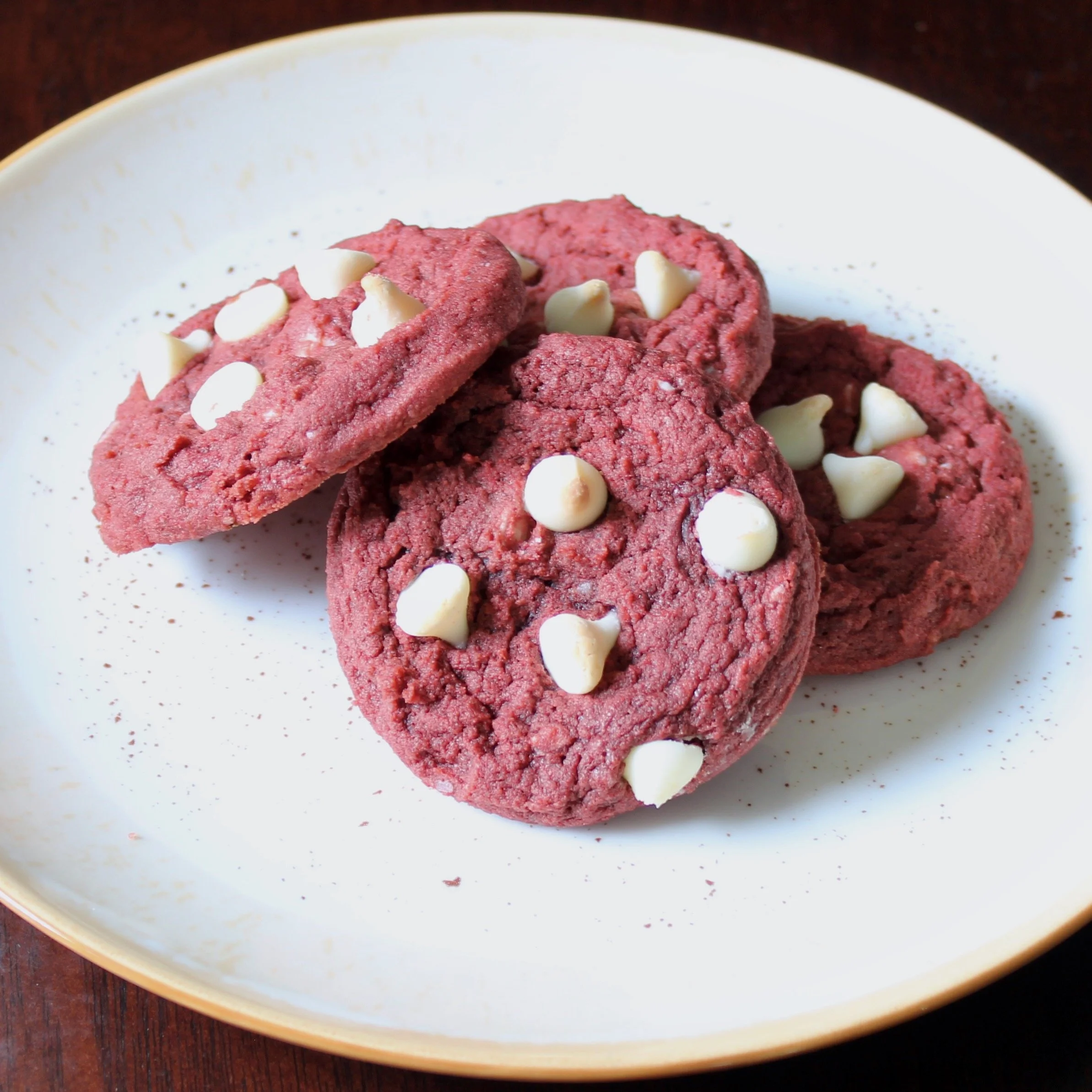 Four red velvet cookies with white chocolate chips stacked together on a white plate over a dark wooden background.