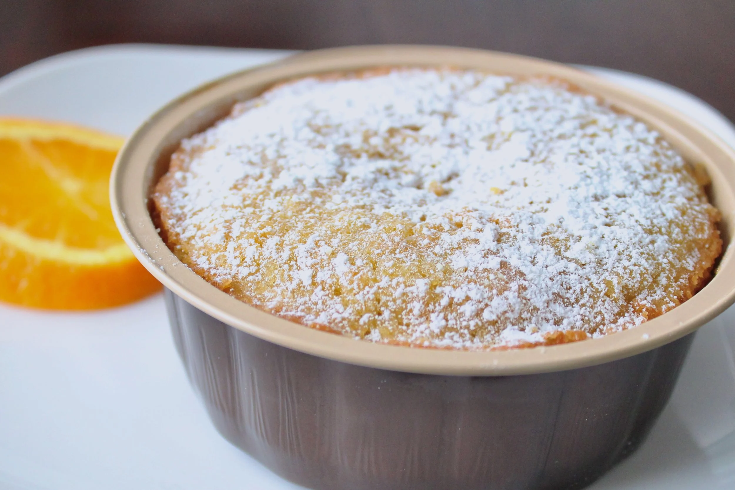 A whole orange olive oil cake in a baking dish, dusted with powdered sugar with a fresh orange slice next to it.