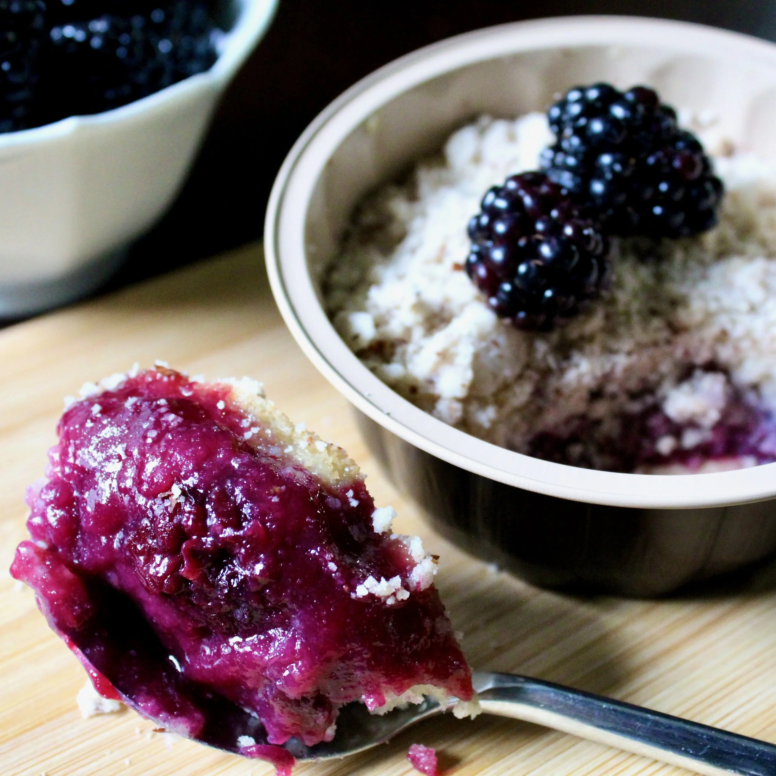 A spoonful of vibrant purple blackberry cobbler filling sits in the foreground next to the dessert bowl.