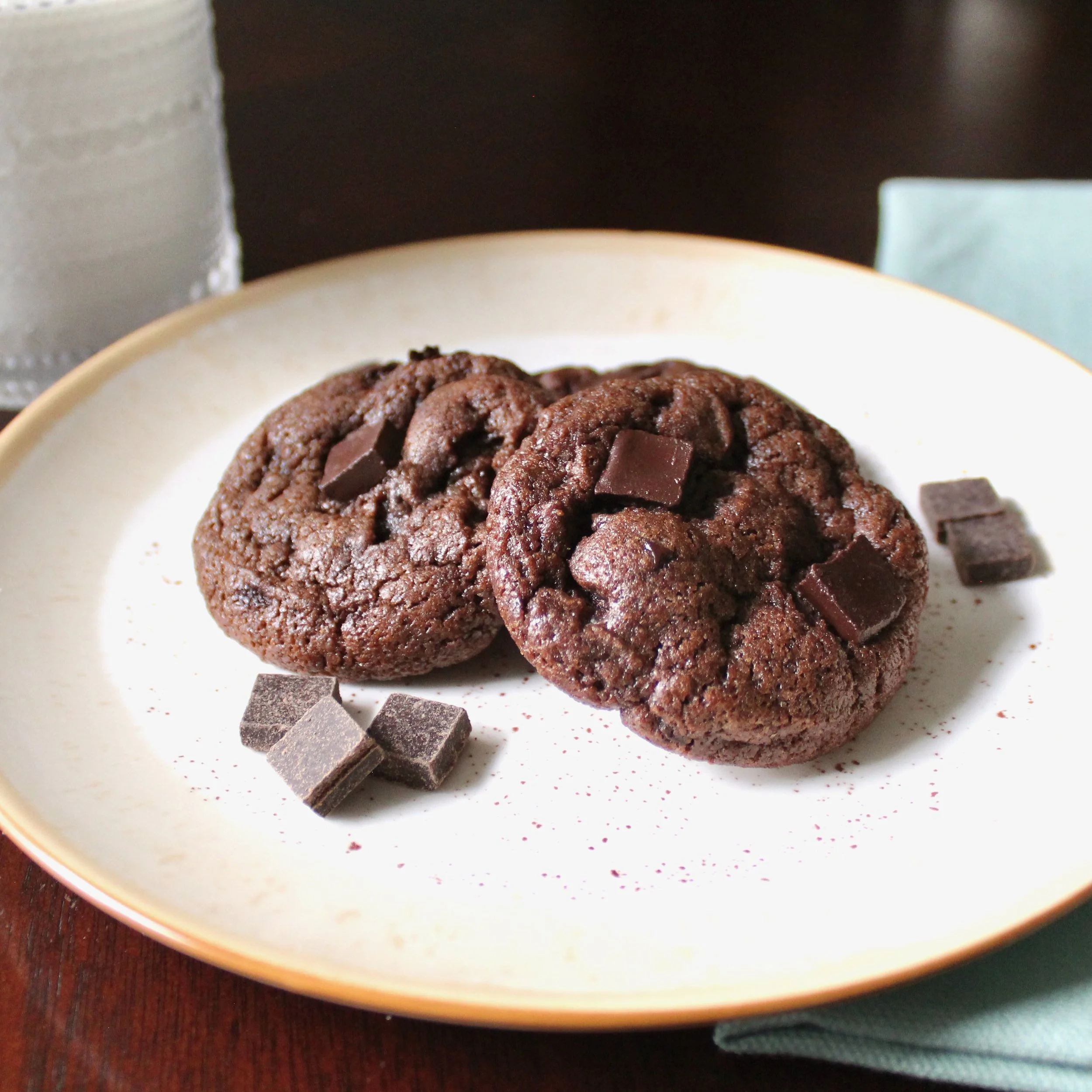 Eye-level shot of thick double chocolate chunk cookies on a plate beside a glass of milk and chocolate chunks.