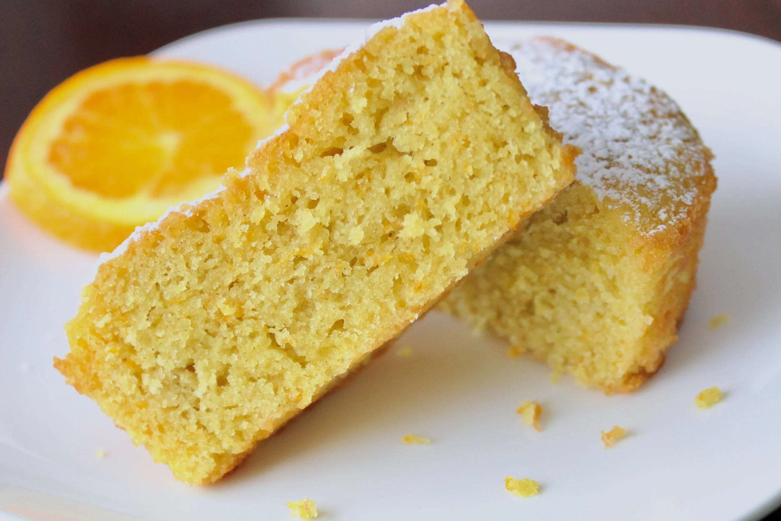 Close-up of a sliced, moist orange olive oil cake dusted with powdered sugar, with a fresh orange slice in the background.