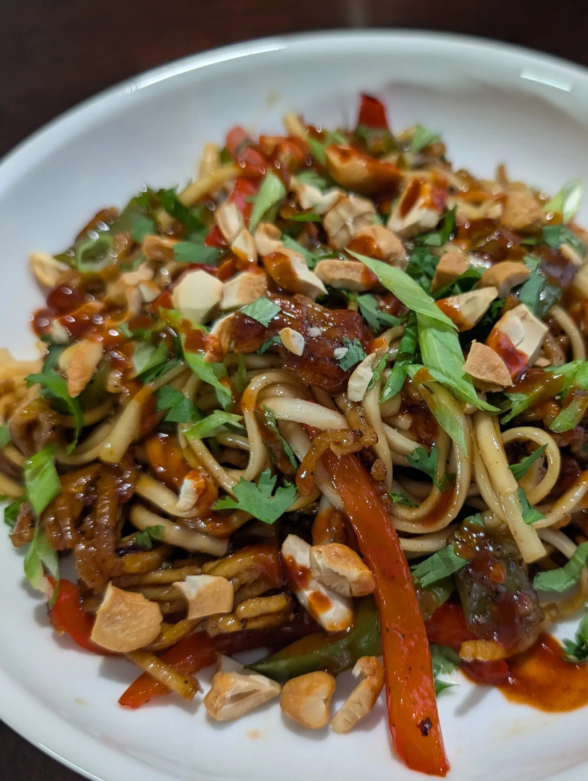 Plate of Asian-style stir-fried noodles with vegetables, chopped nuts, cilantro, and sauce.