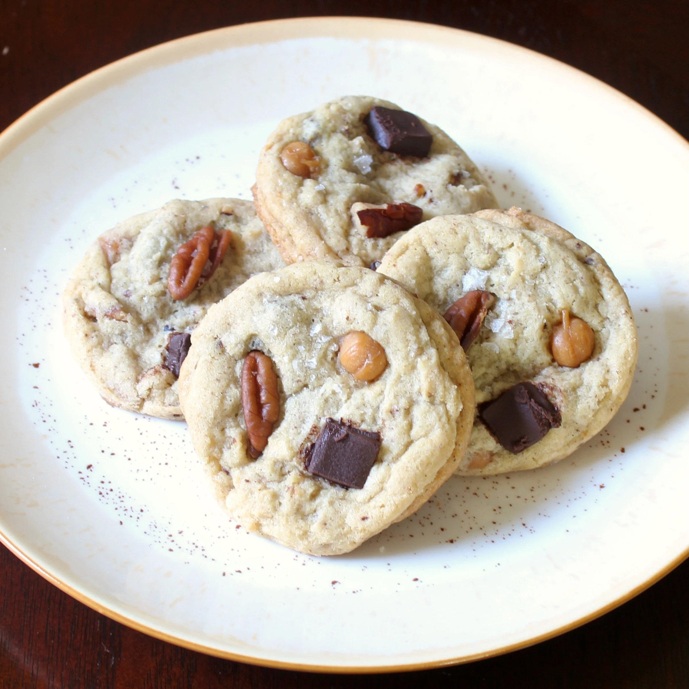 Four pecan and chocolate chunk cookies stacked on a round cream plate over a dark wood table.