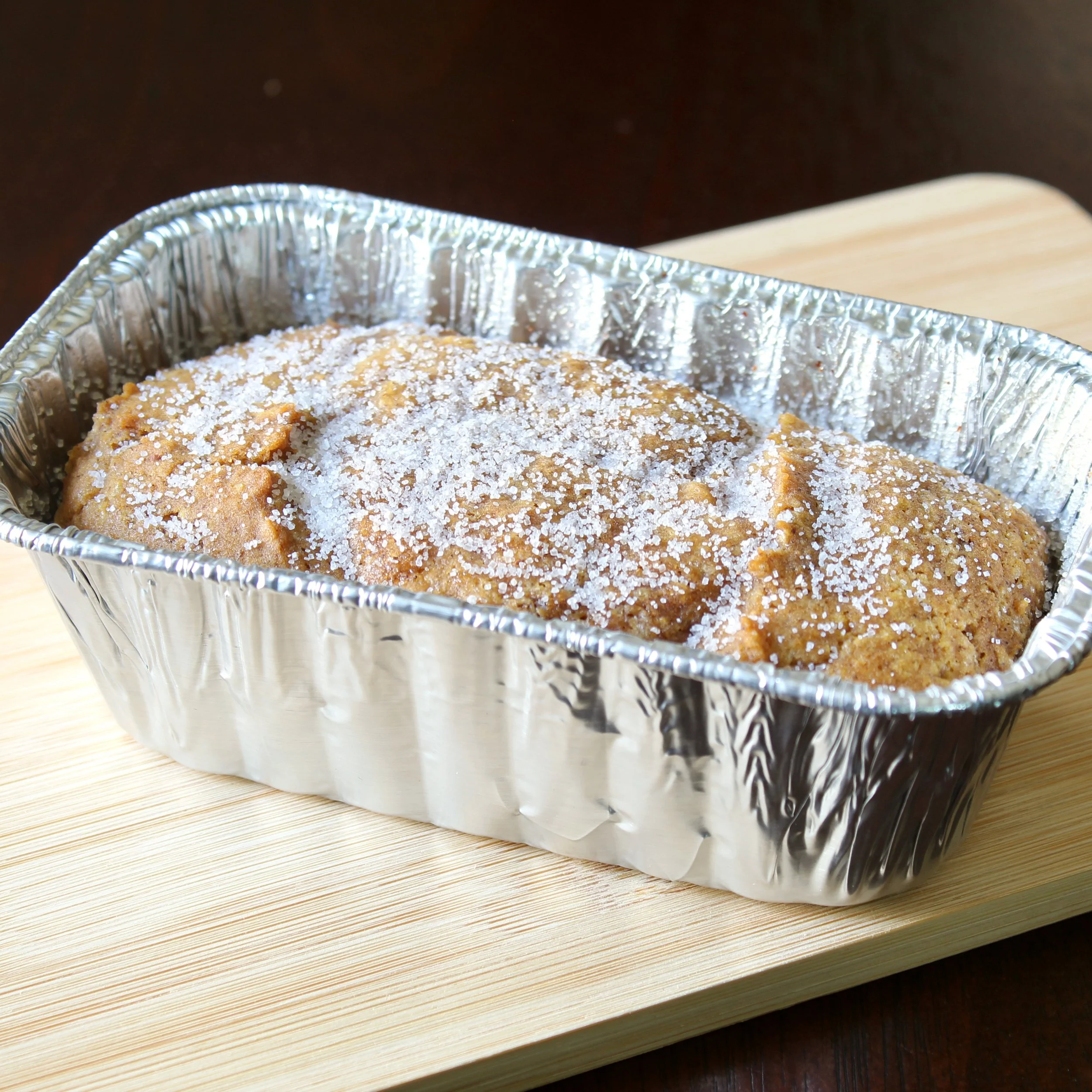 Sugar-topped pumpkin bread loaf baked in an aluminum foil pan on a bamboo board.