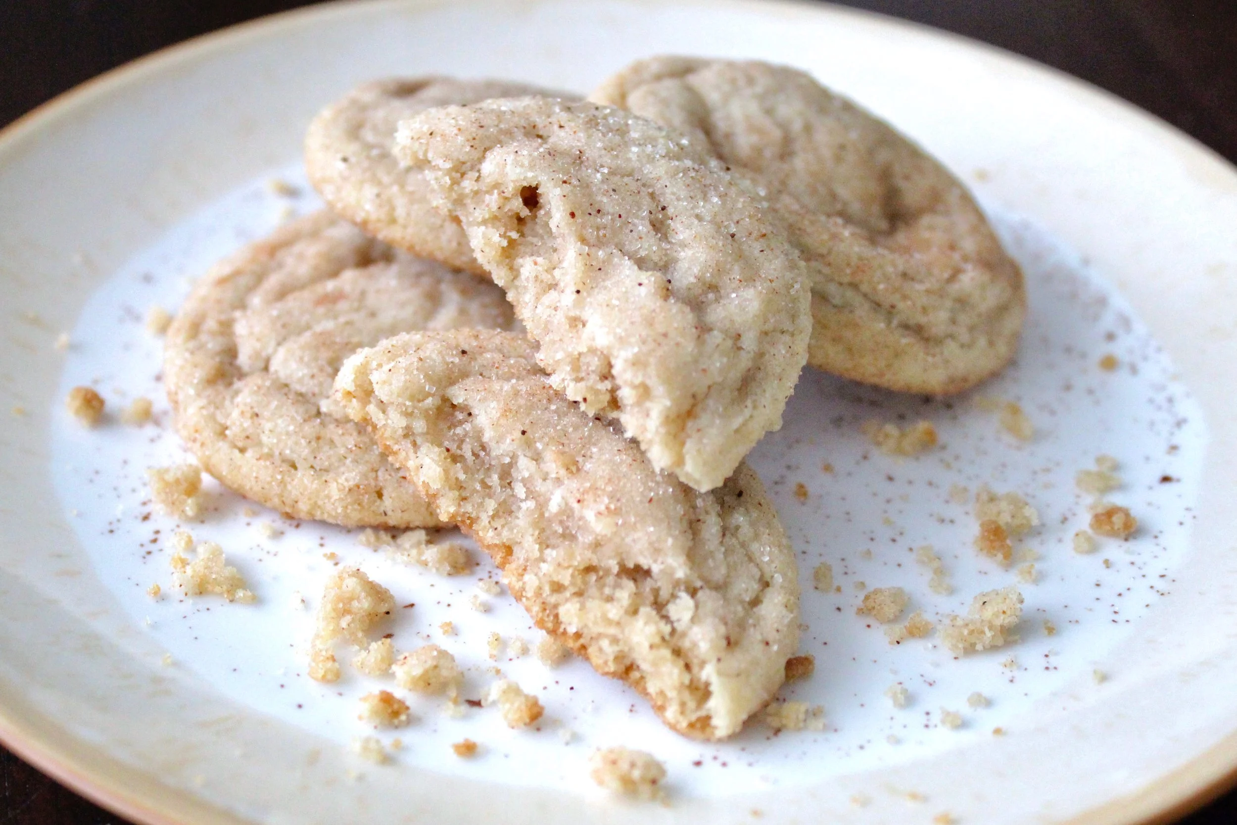 A broken snickerdoodle cookie stacked on others on a white plate, showing a soft texture and cinnamon sugar coating.