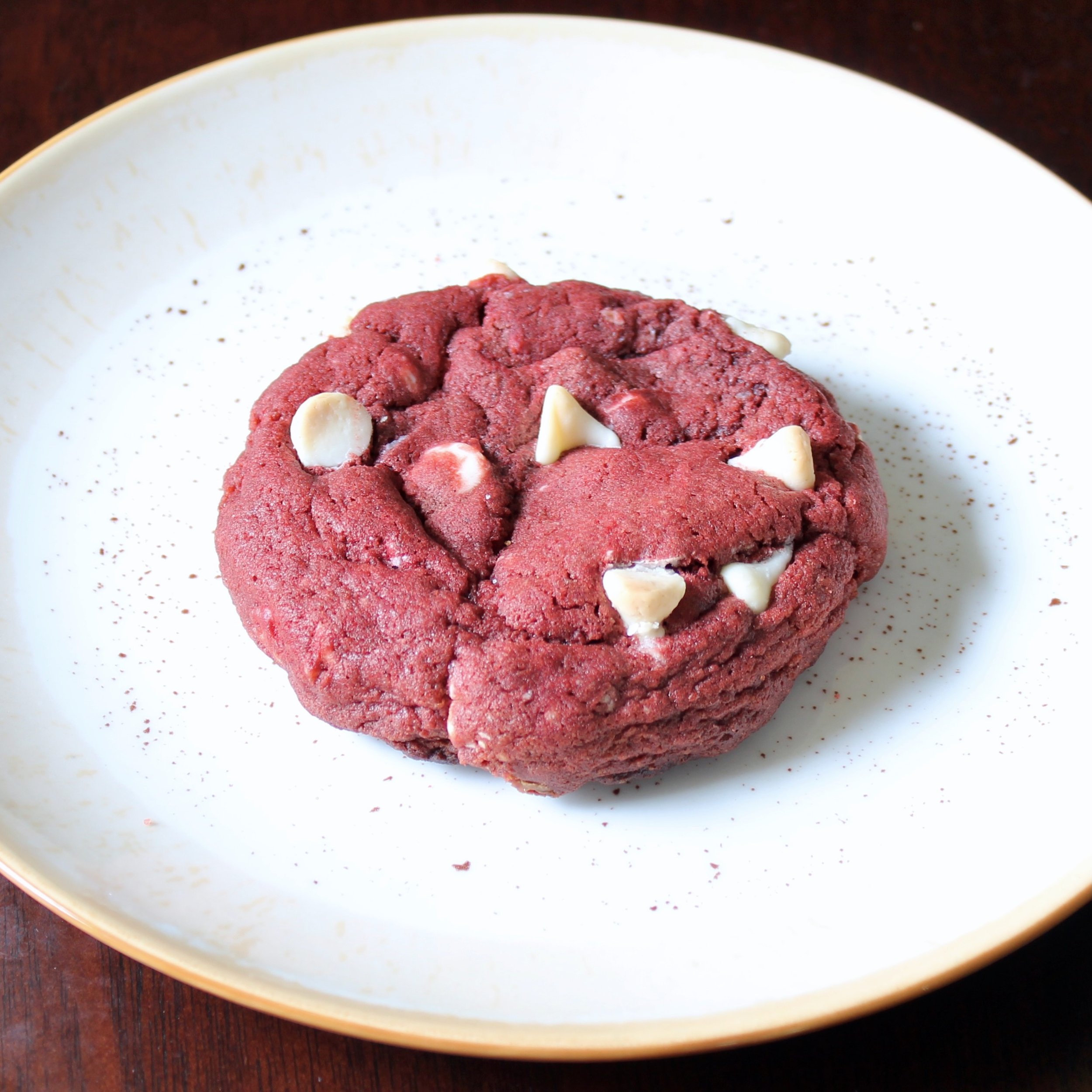 Single thick red velvet cookie studded with white chocolate chips sitting centered on a white speckled plate.
