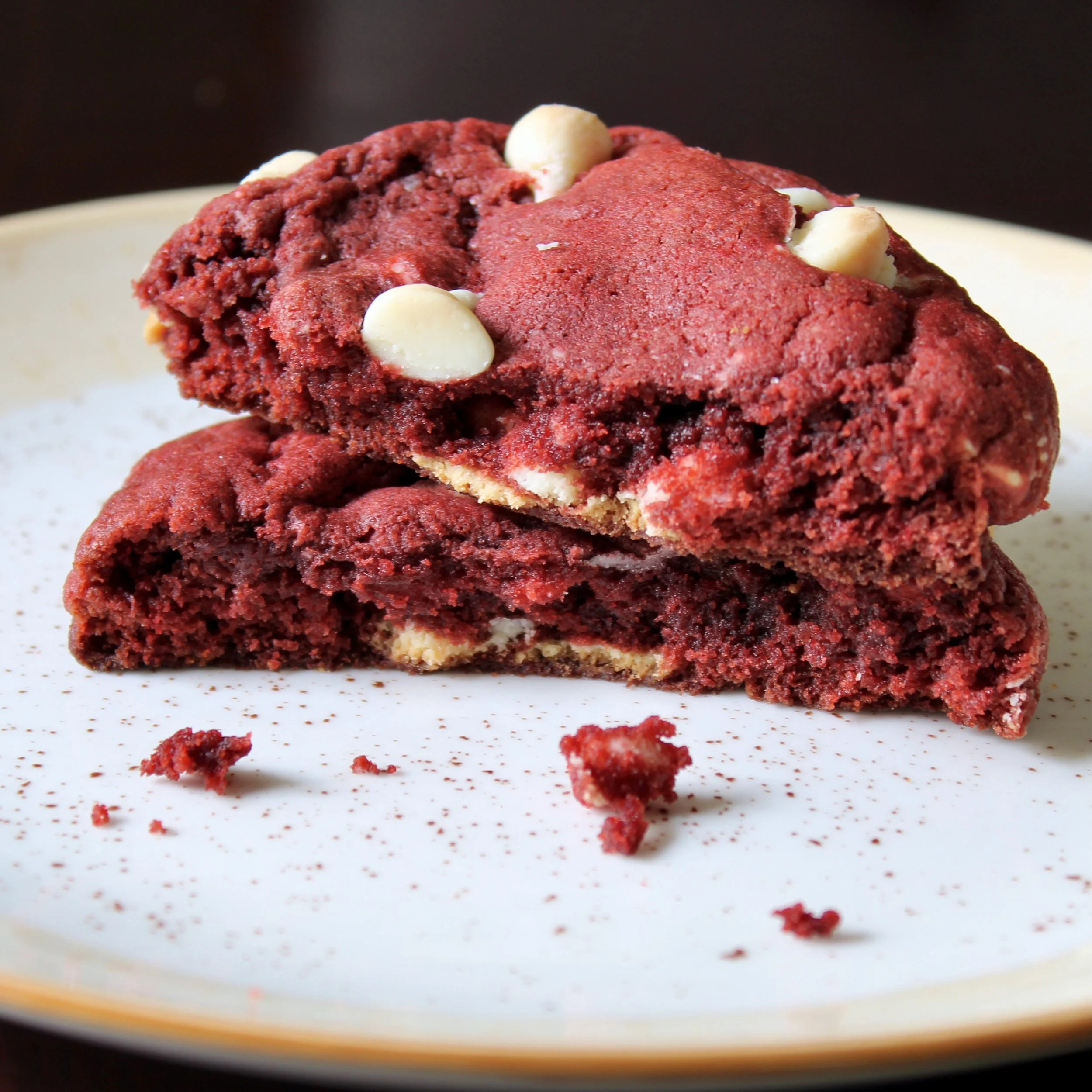 Thick red velvet cookie broken in half, stacked on a plate to show the chewy center and white chocolate chips.