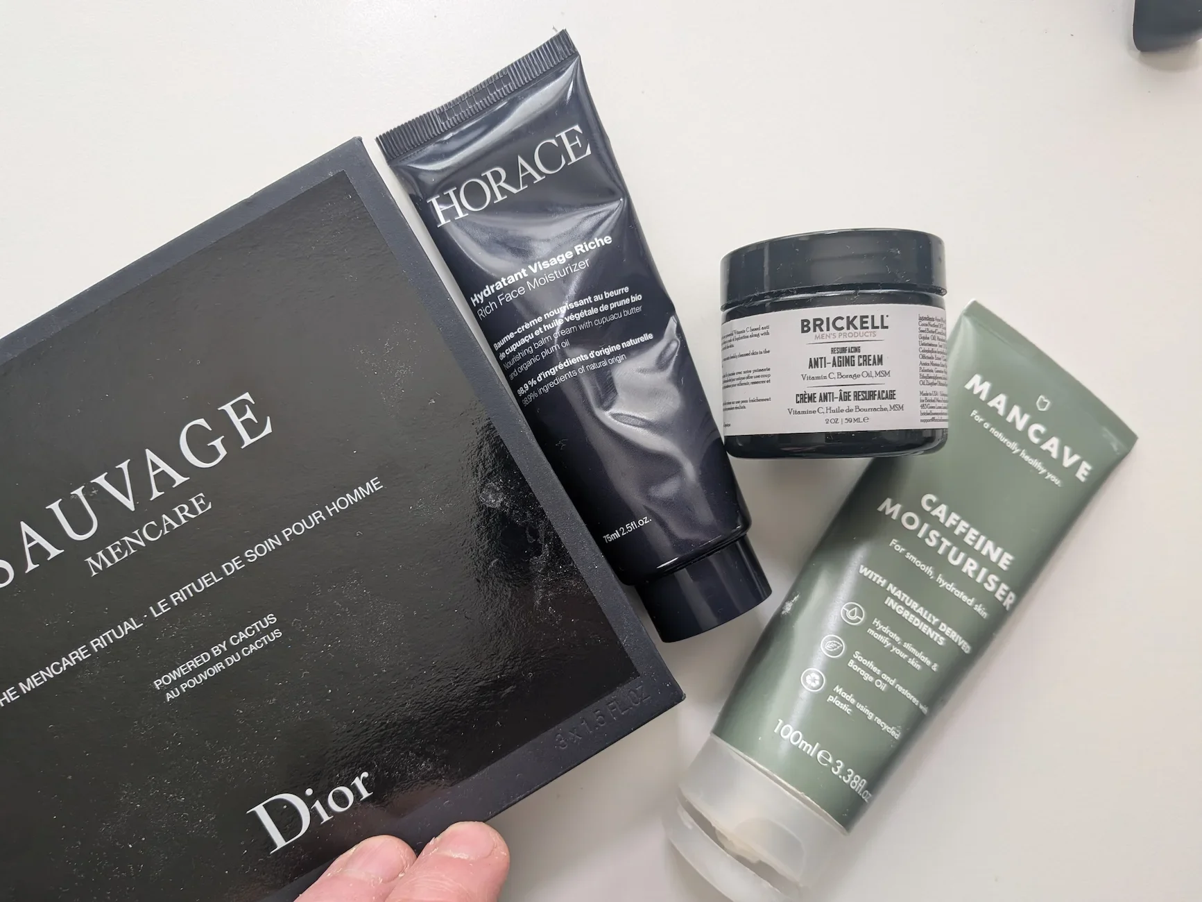 A top-down view of a curated men's skincare routine featuring Dior Sauvage Mencare, Horace Rich Face Moisturiser, Brickell Anti-Aging Cream, and ManCave Caffeine Moisturiser.