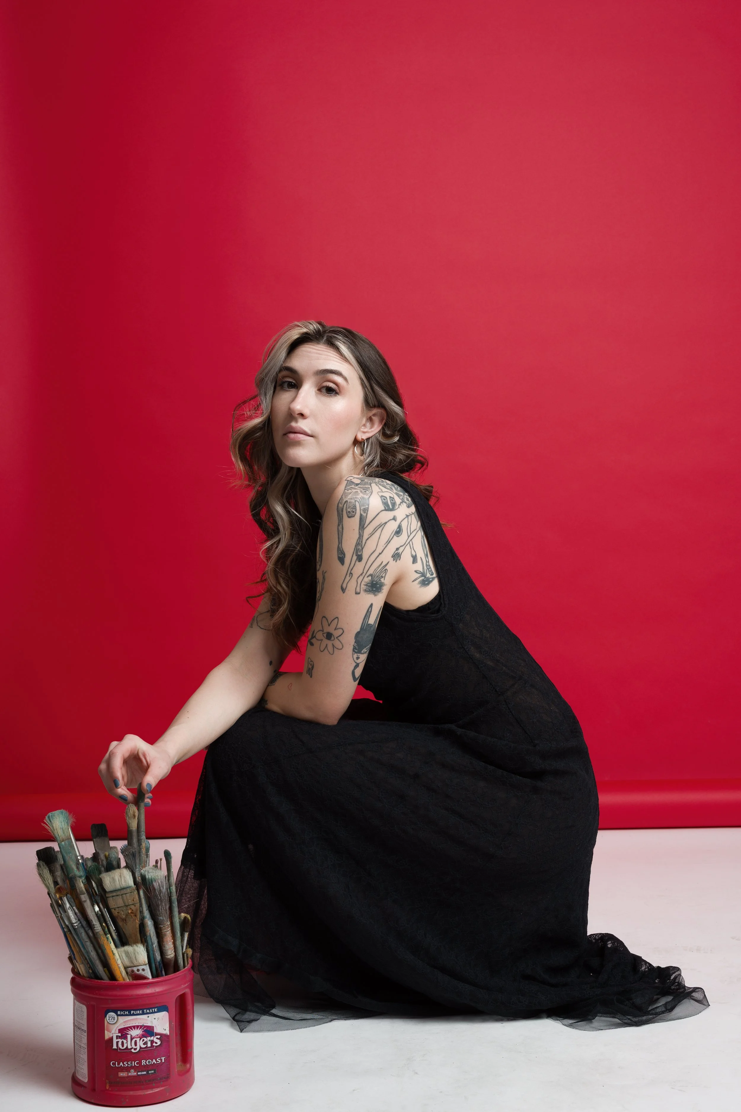 A young woman with wavy hair, tattoos on her left arm, wearing a black dress, sitting on the floor in front of a red backdrop, holding a container of paintbrushes.
