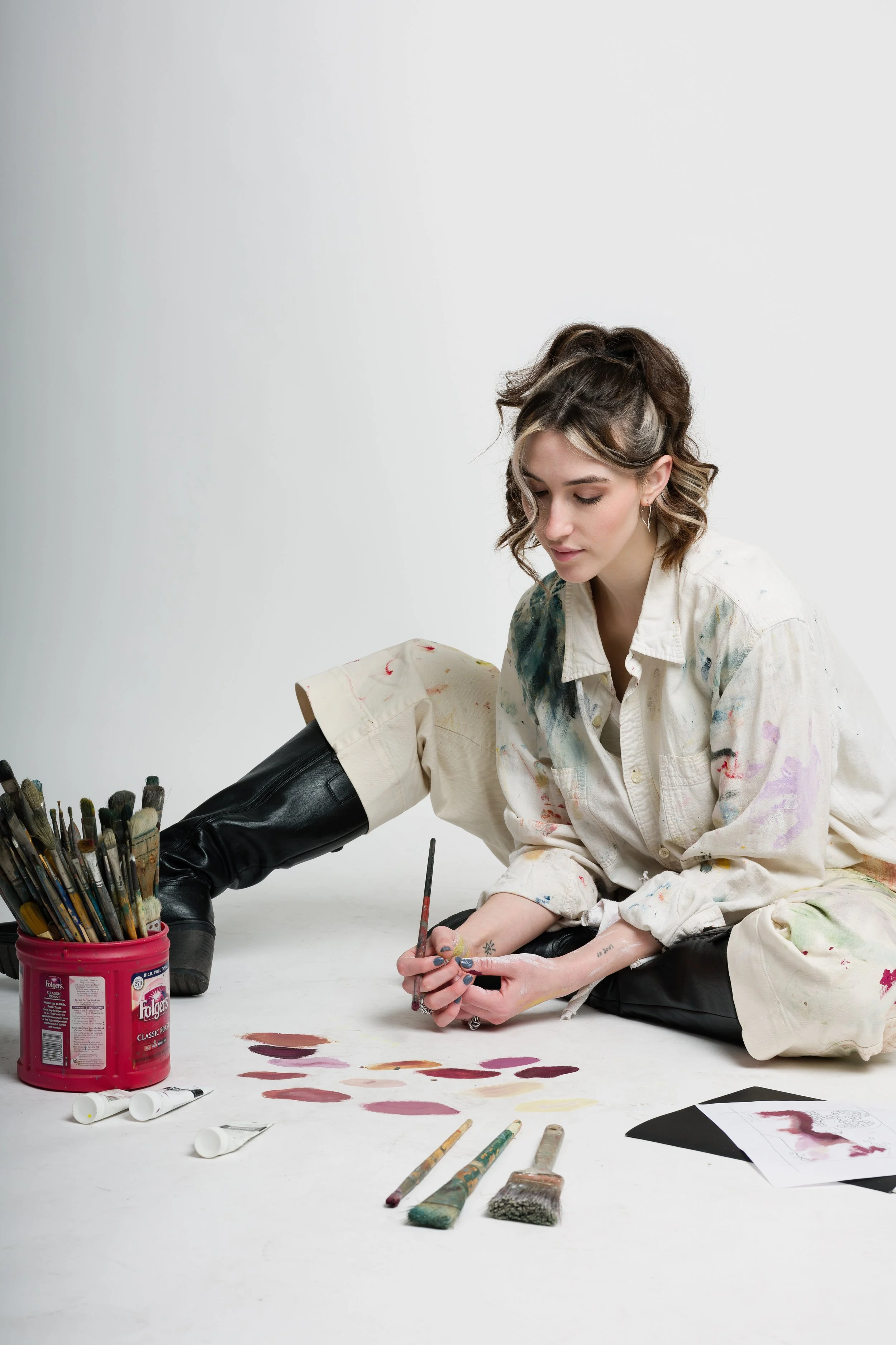 Young woman creating abstract art on the floor with paintbrushes, paints, and tubes of paint around her, wearing a paint-splattered shirt and black leather boots.