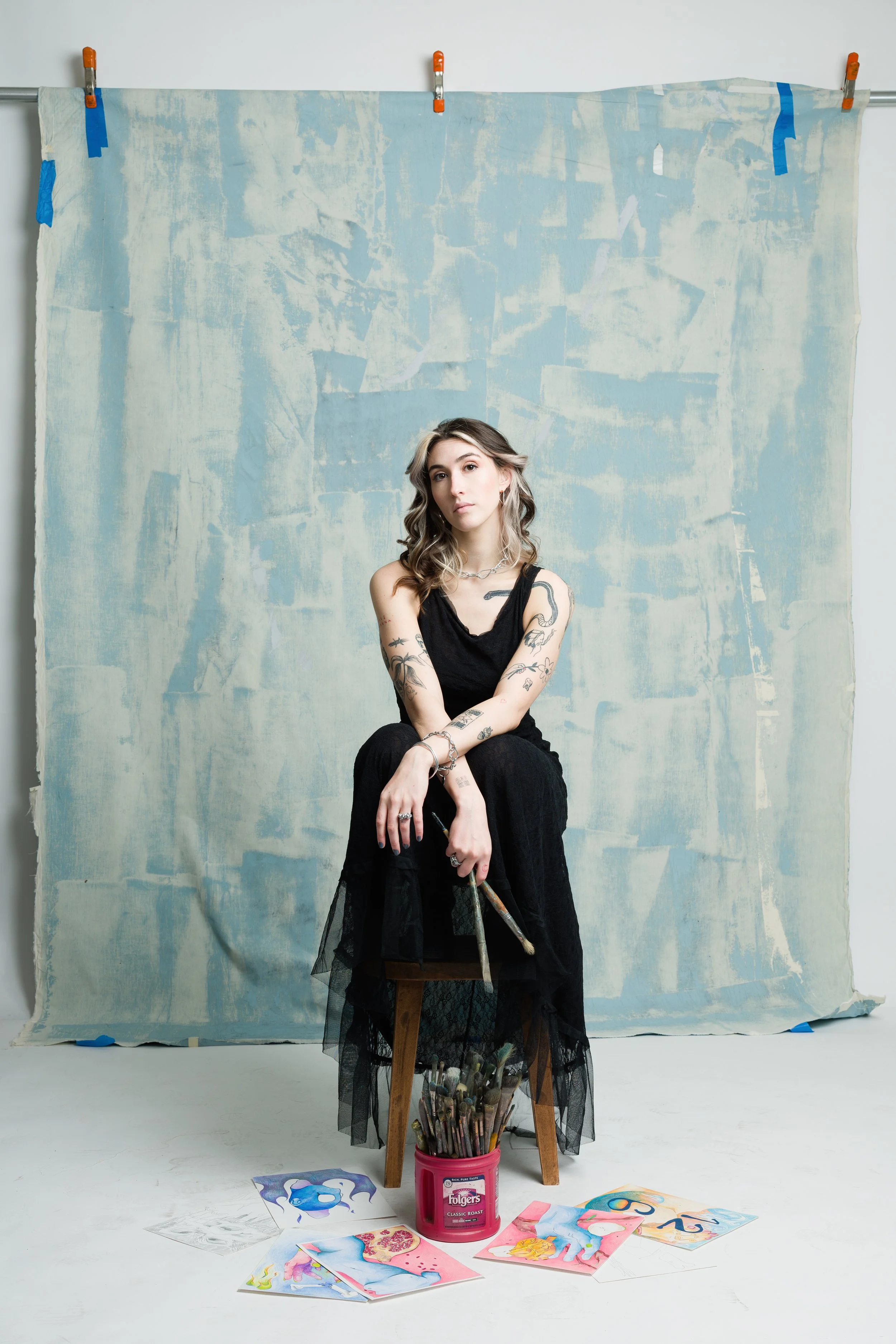 A woman with tattoos sits on a stool in front of a textured blue backdrop, surrounded by colorful artwork and brushes.