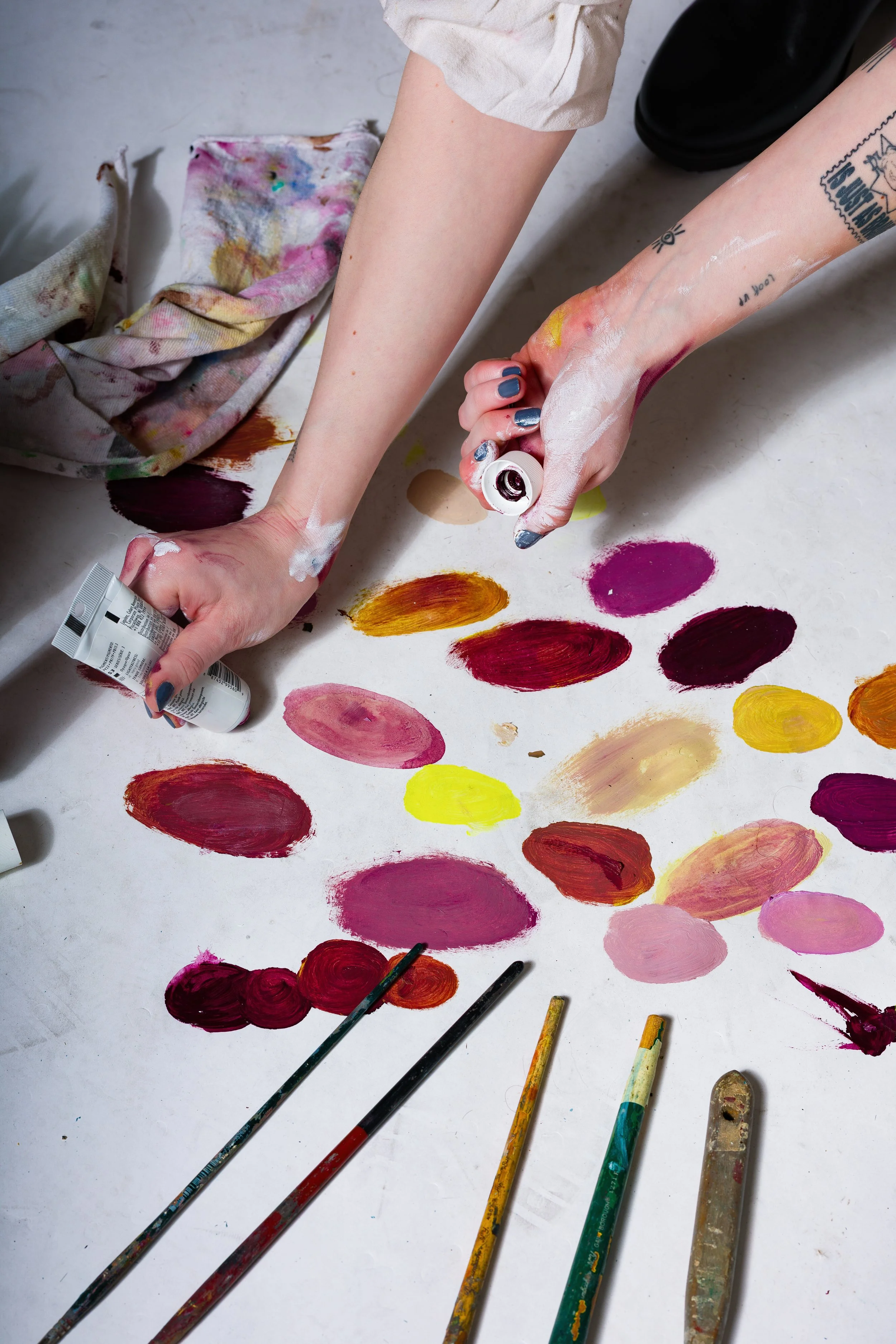 Person's hands mixing colorful paint on a white surface, surrounded by paintbrushes and palettes.