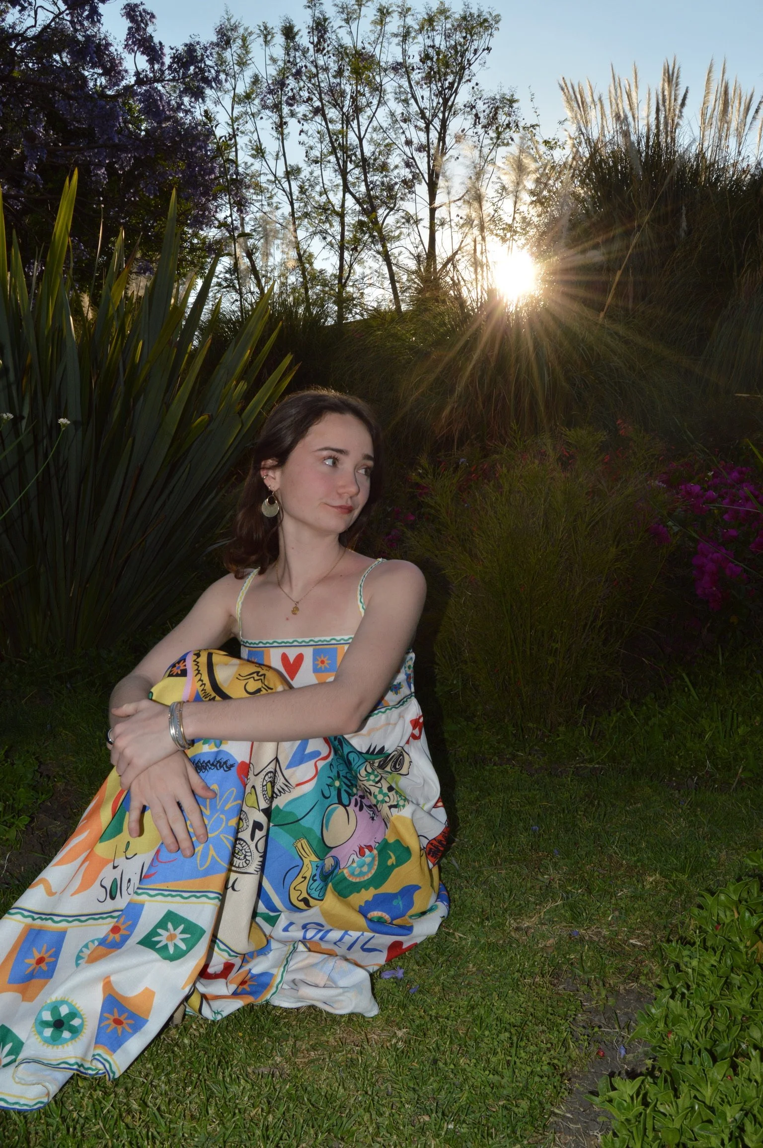 A young woman sitting on grass in a garden during sunset, wearing a colorful, patterned dress and earrings, with trees and sunlight in the background.