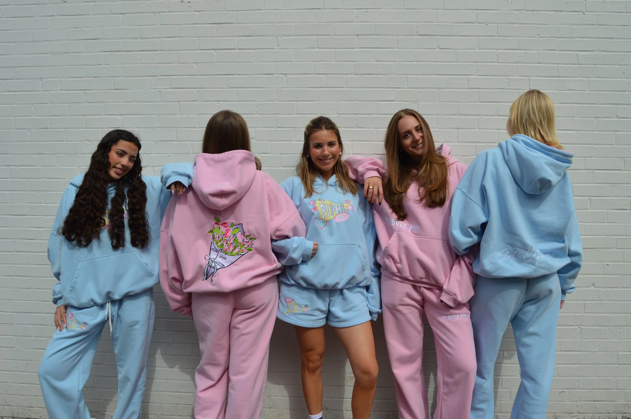 Group of five young women wearing pastel blue and pink hoodies and shorts, posing against a brick wall. One woman faces backward, the other four face forward, smiling.