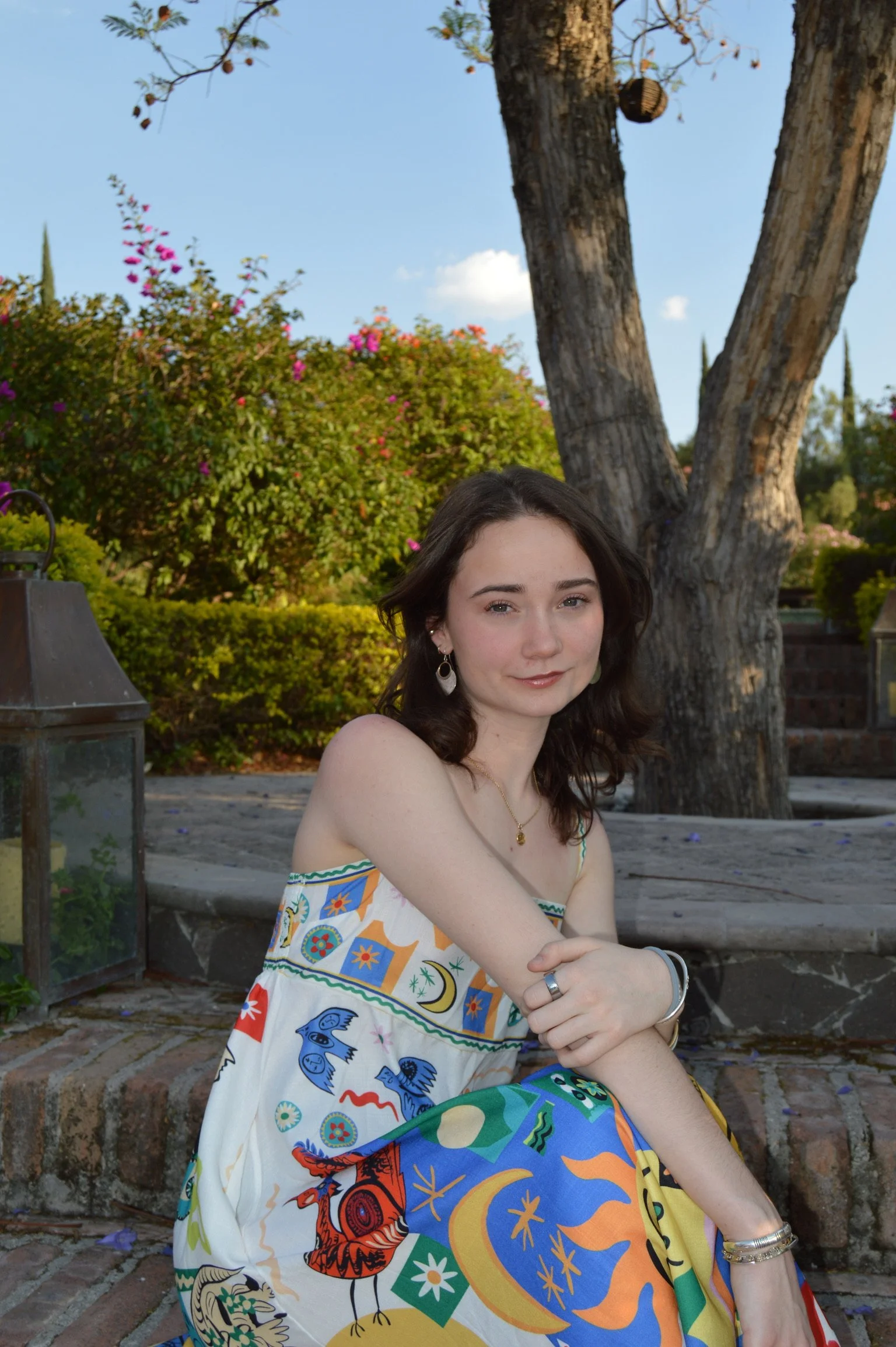 Young woman with dark hair wearing a colorful strapless dress, sitting outdoors near a tree, with gardening lanterns and flowering bushes in the background.