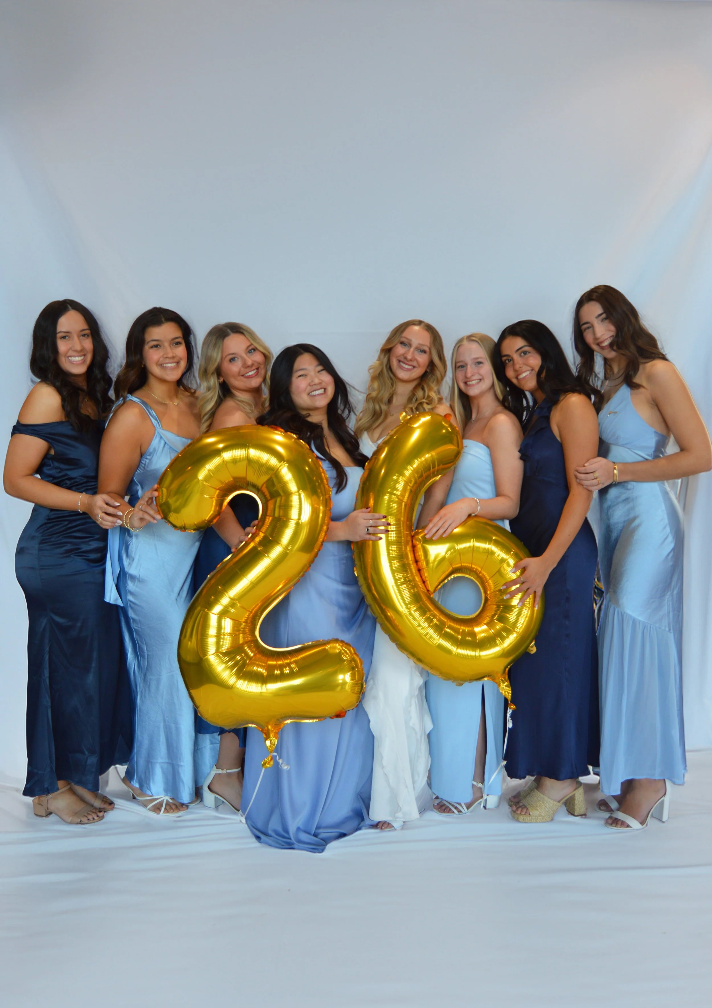Group of nine women celebrating 26th birthday, holding gold balloons shaped as '2' and '6' against a neutral background, dressed in blue and white dresses.