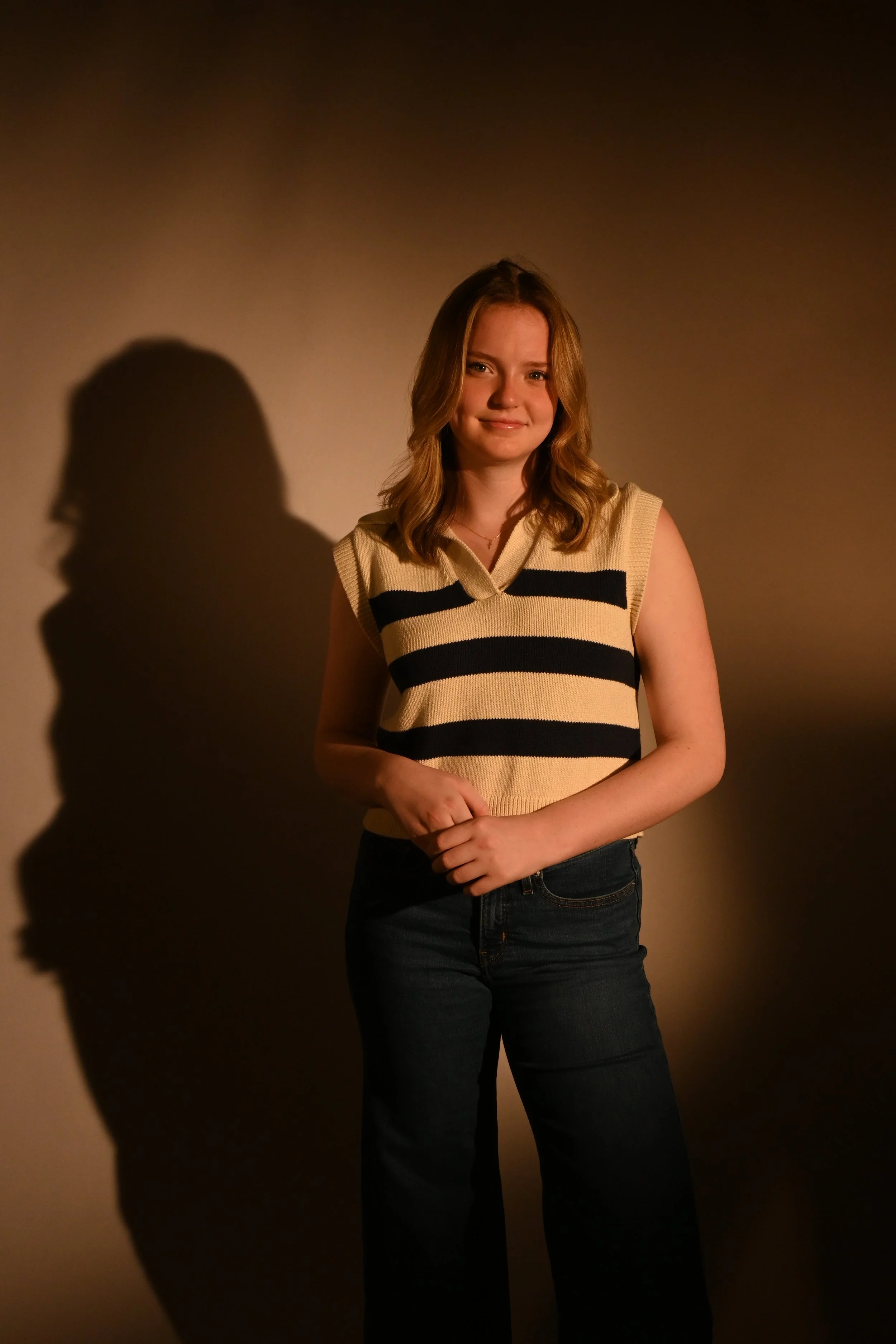 A young woman standing in front of a plain wall with a shadow cast beside her. She is smiling slightly, wearing a sleeveless, striped sweater and dark jeans.