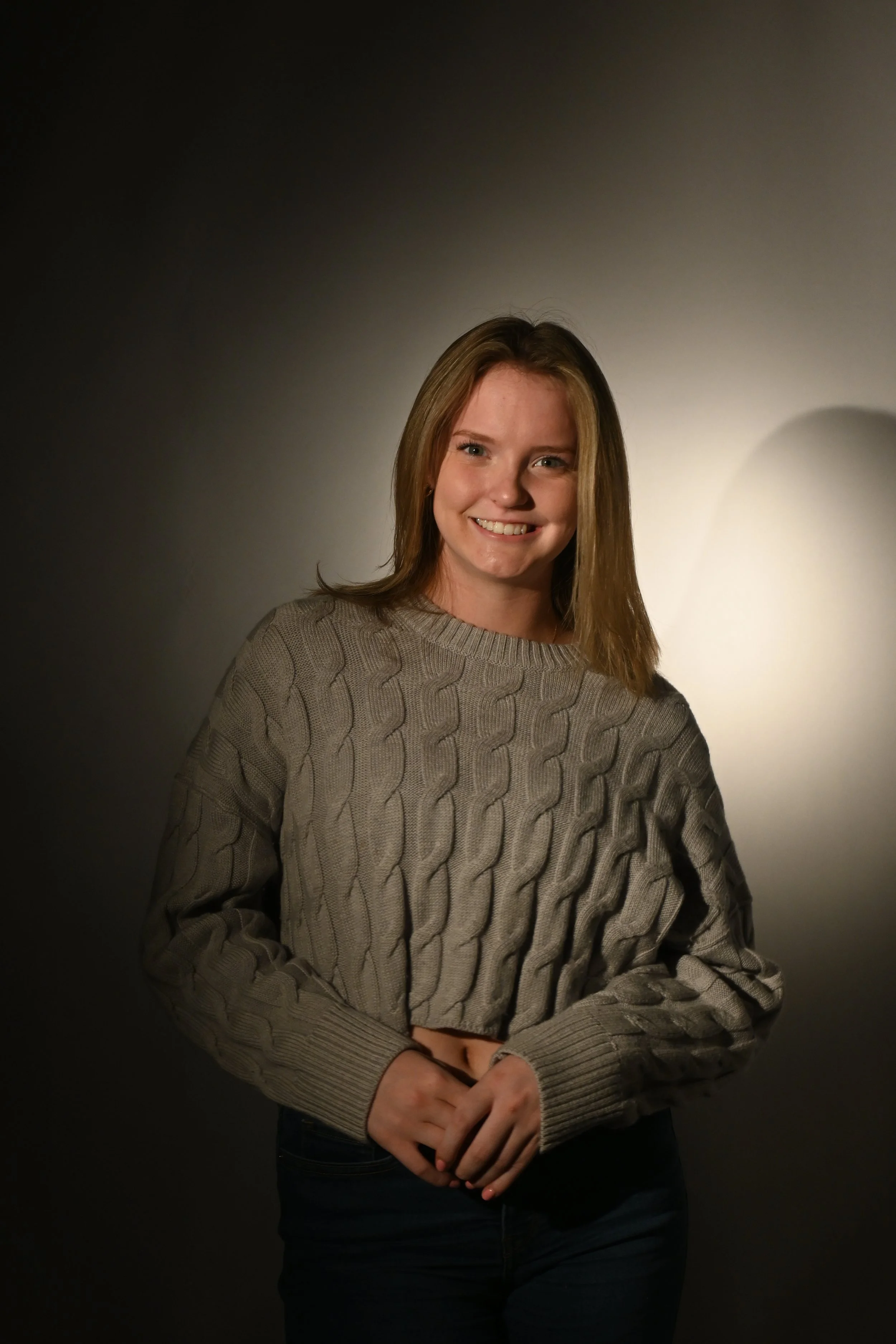 Young woman with light brown hair smiling, wearing a gray cable-knit sweater, standing against a dark background with a soft light illuminating her face.