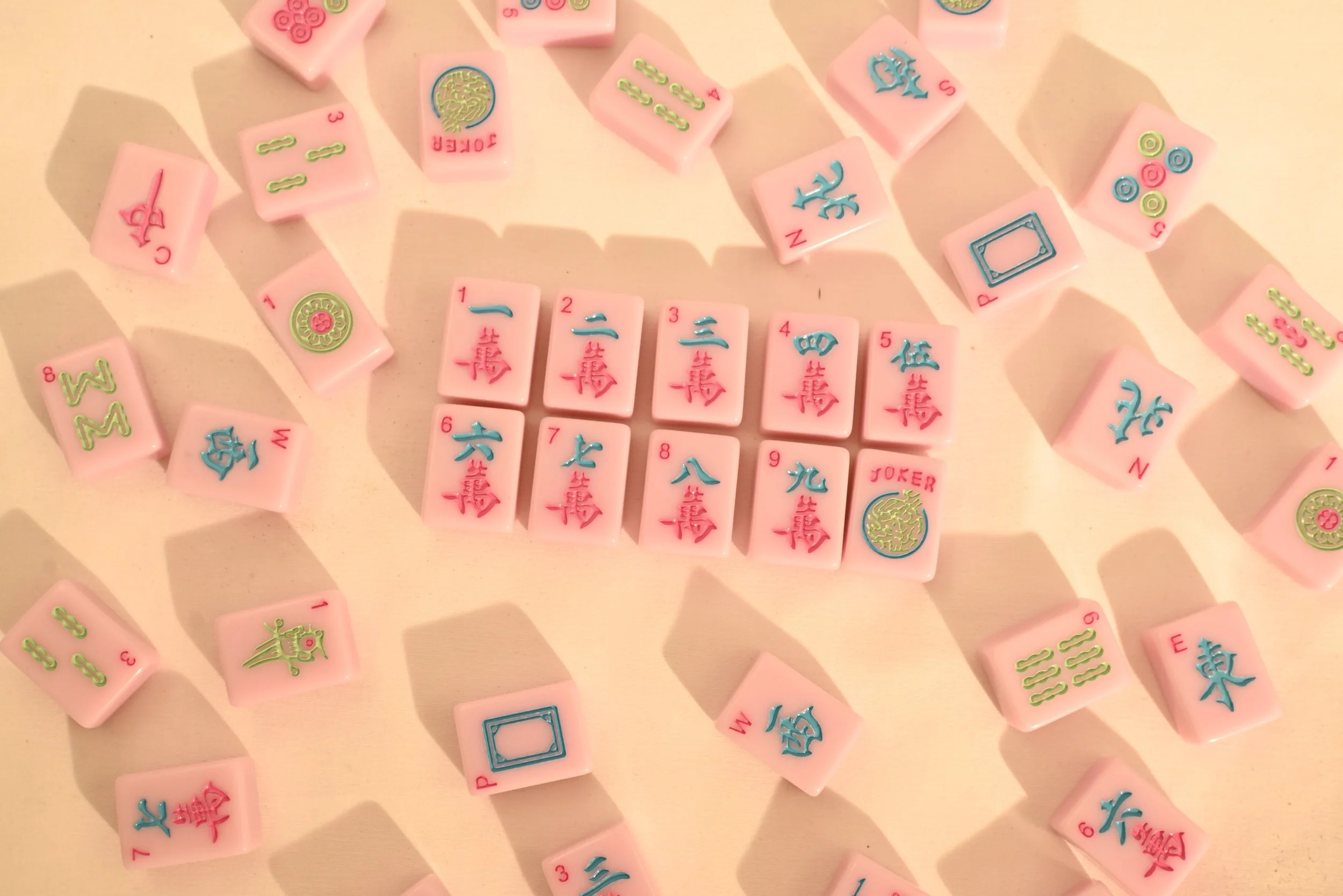 Pink Mahjong tiles with blue and red Chinese characters, numbers, and symbols, laid out on a light-colored surface. Some tiles form a sequence in the center, with scattered tiles around.