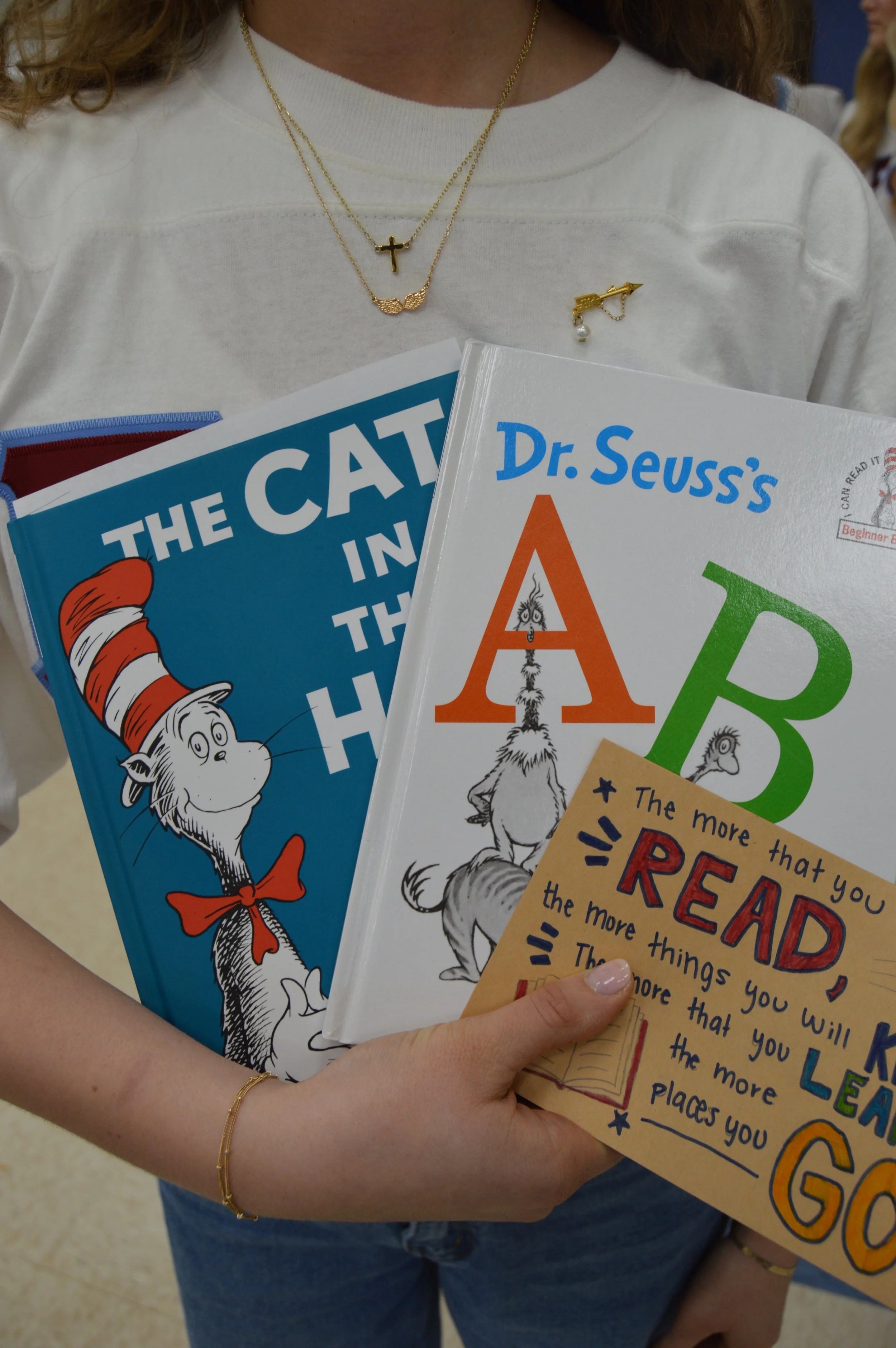 Person holding multiple children's books, including "The Cat in the Hat," "Dr. Seuss's ABC," and a reading motivation card, with necklaces and a pin on their white shirt.