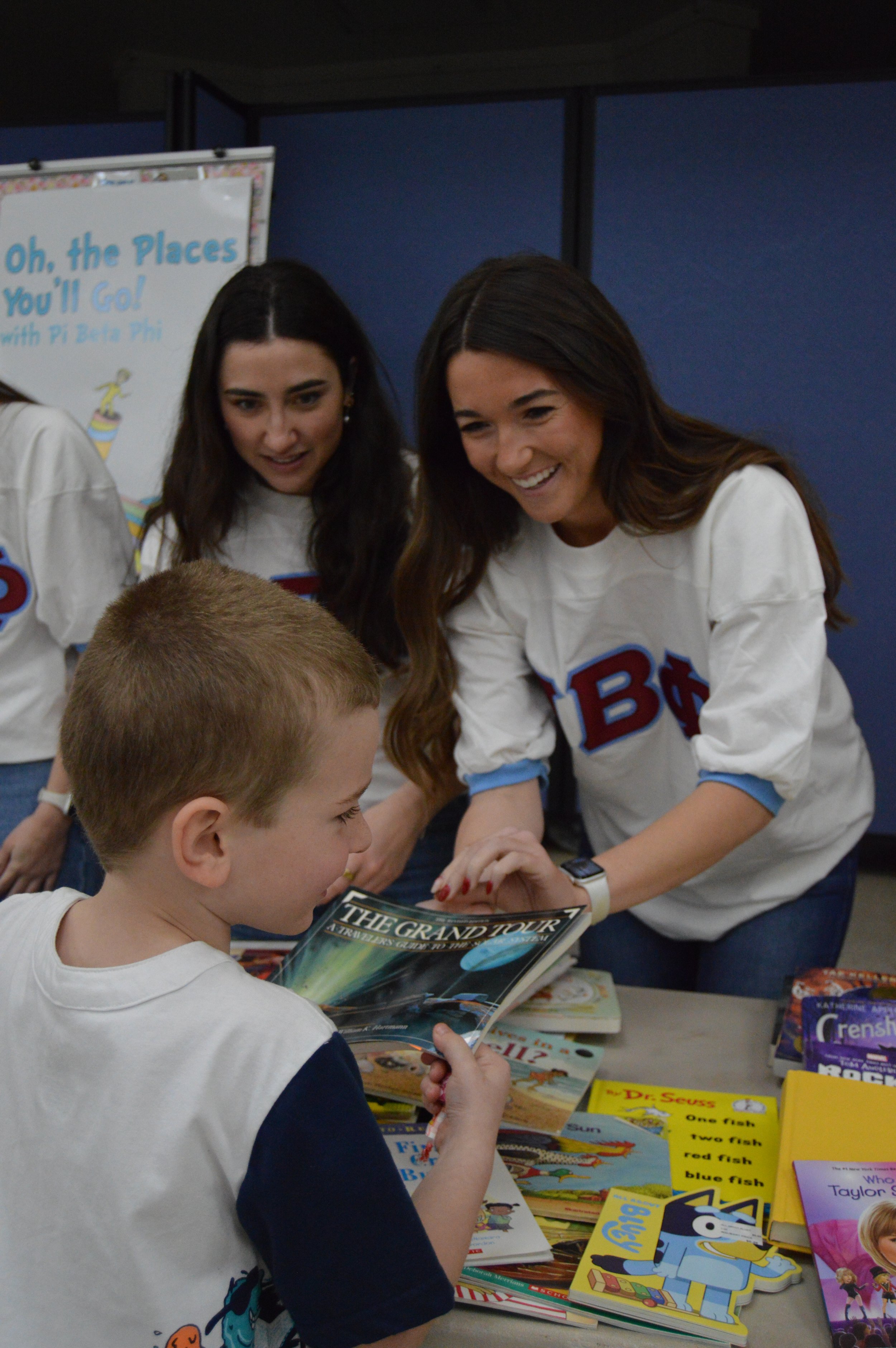 Two women in white shirts are smiling while showing children's books to a young boy who is holding a book and smiling. Books and a poster are on a table in front of them.