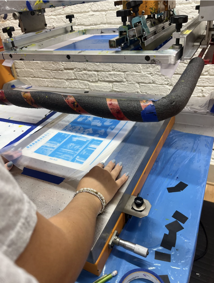 A person operating a screen printing machine to produce a blue-colored print on a sheet of paper or plastic, with their hand near the printed material and black tape on the blue surface.
