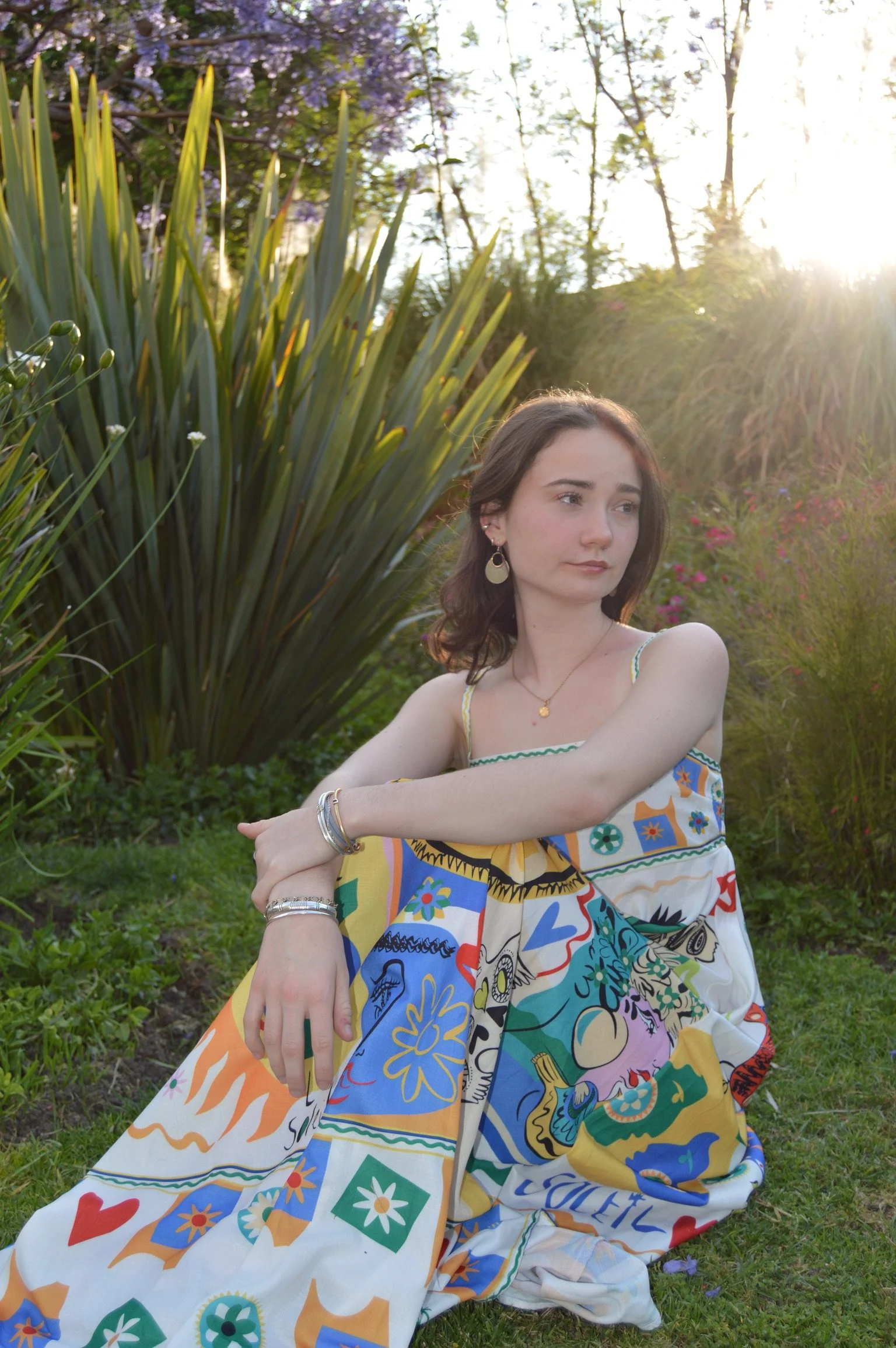 A young woman sitting outdoors among green plants and flowers, with sunlight shining in the background, wearing a colorful patterned dress and jewelry.