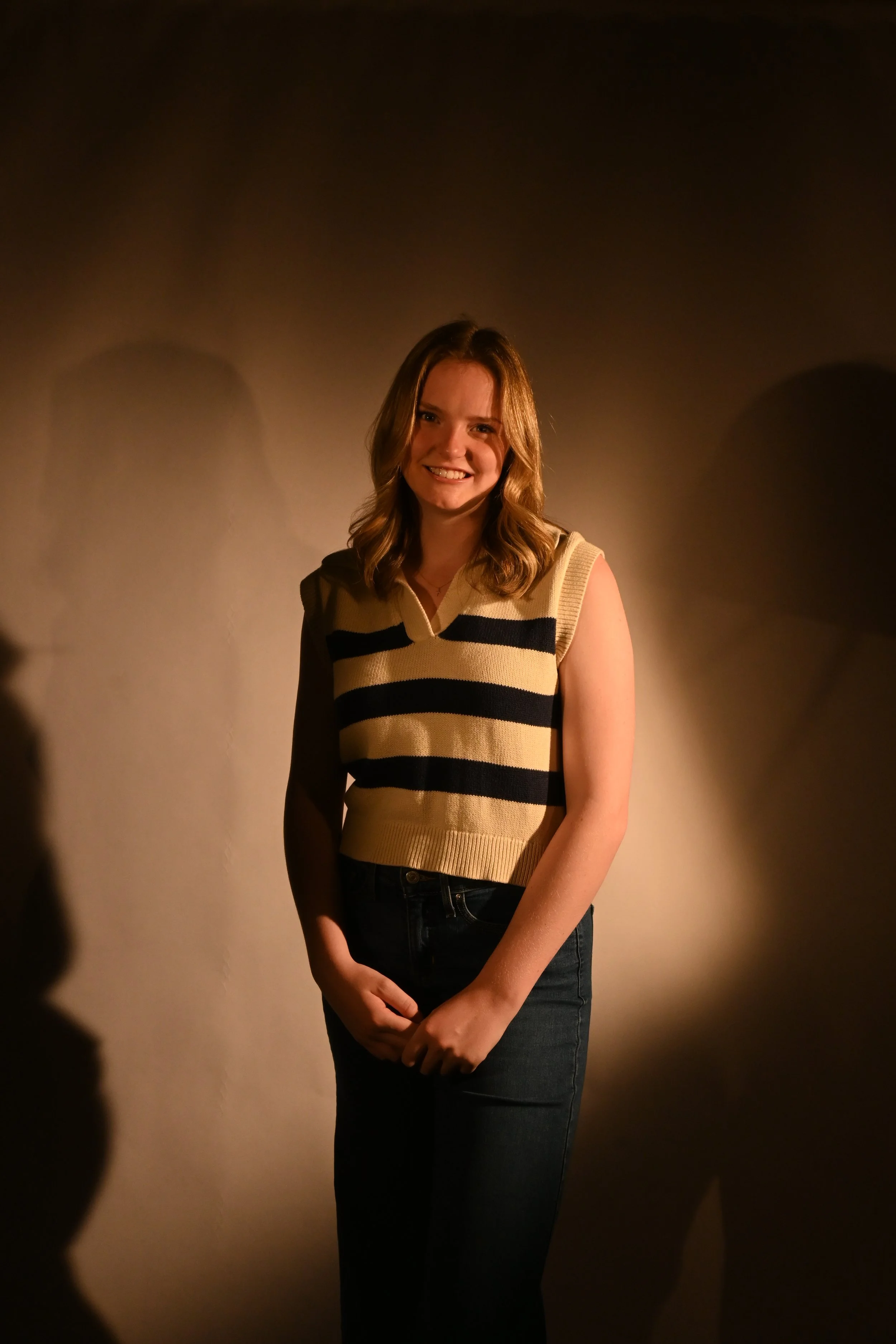 A young woman with shoulder-length brown hair smiling and standing against a beige wall with her hands clasped in front of her. She is wearing a sleeveless yellow and black striped sweater and dark jeans.