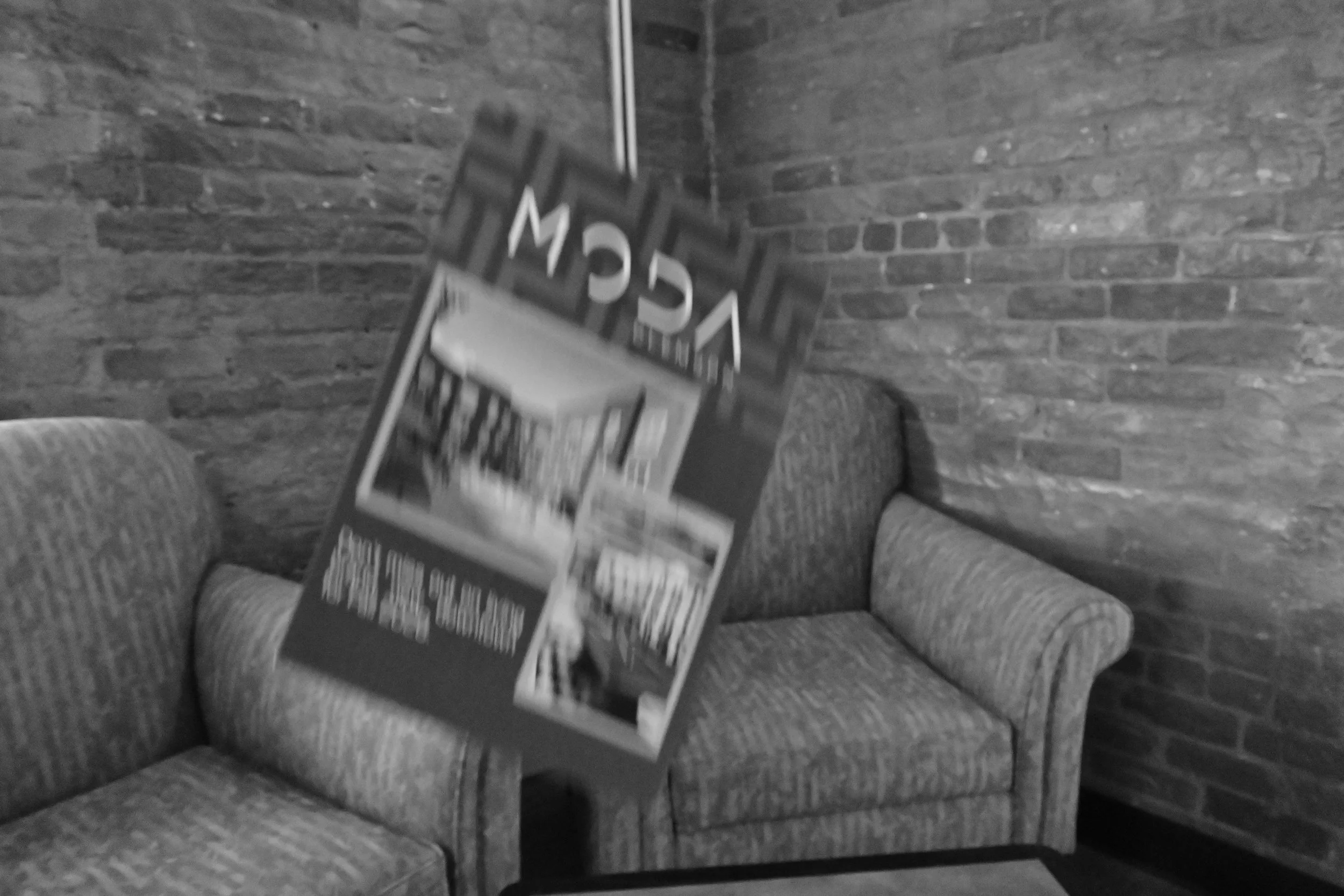 A black and white photo of a waiting area with a fabric upholstered couch against a brick wall. A magazine or brochure is standing on a small table in front of the couch, displaying to the camera, but the text and images on it are blurry and not discernible.