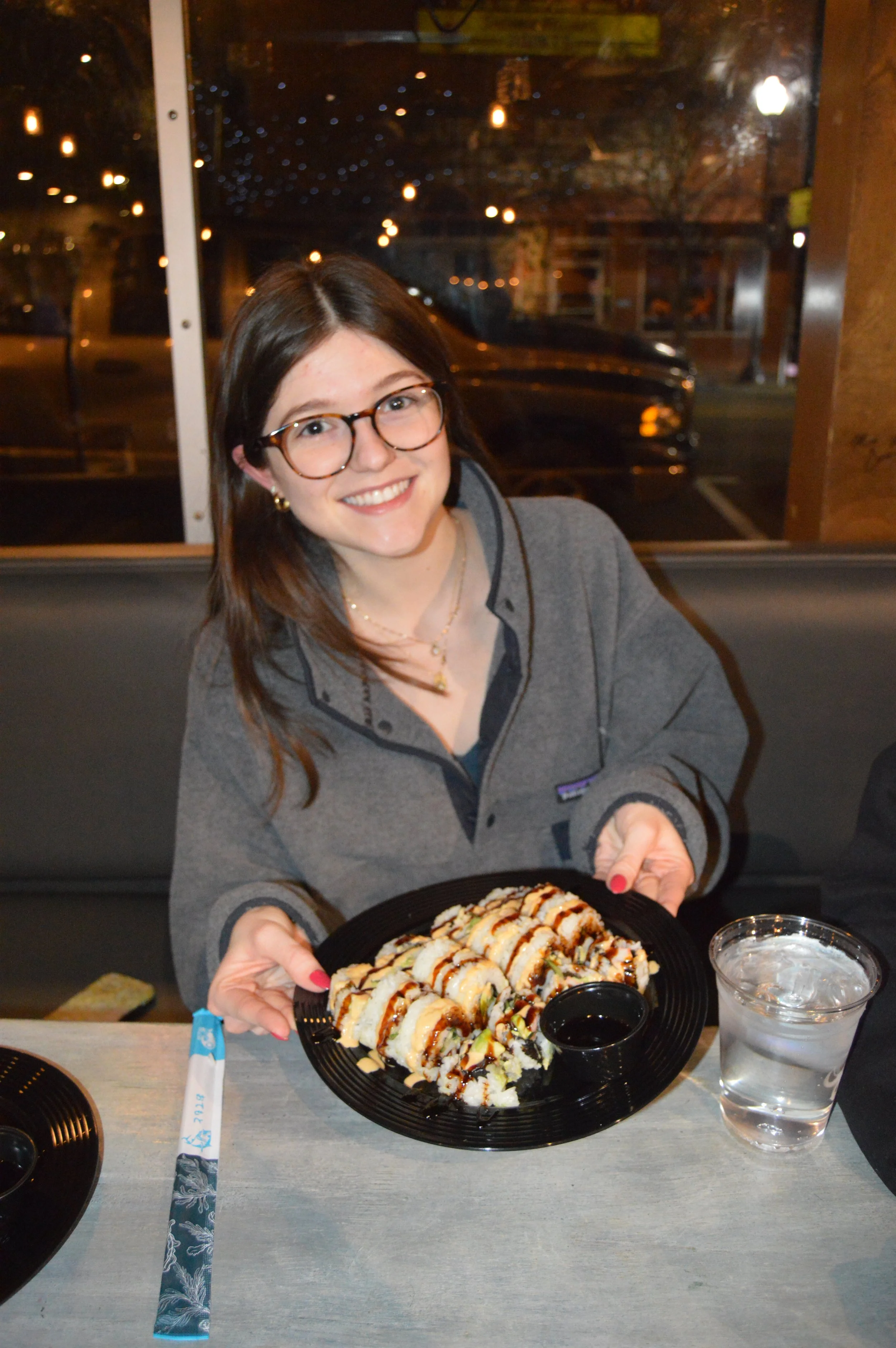 A woman with glasses and a gray jacket holding a plate of sushi with a small container of soy sauce, sitting at a table with a glass of water, in a restaurant at night.
