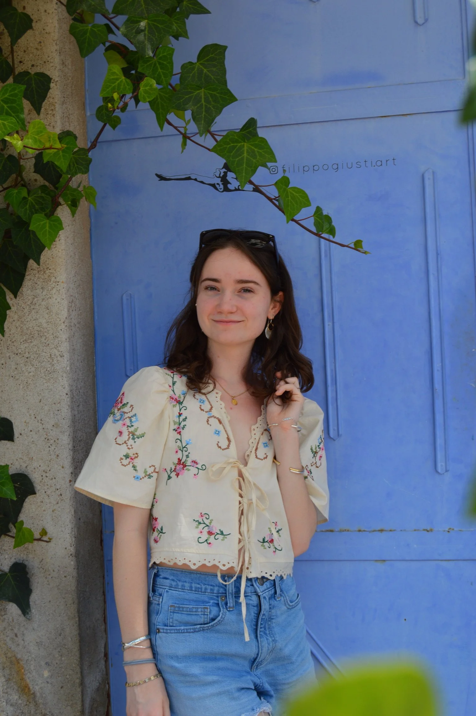 A young woman standing in front of a blue door with climbing green ivy leaves above her. She is smiling, wearing a cream embroidered top, light blue jeans, and sunglasses on her head.