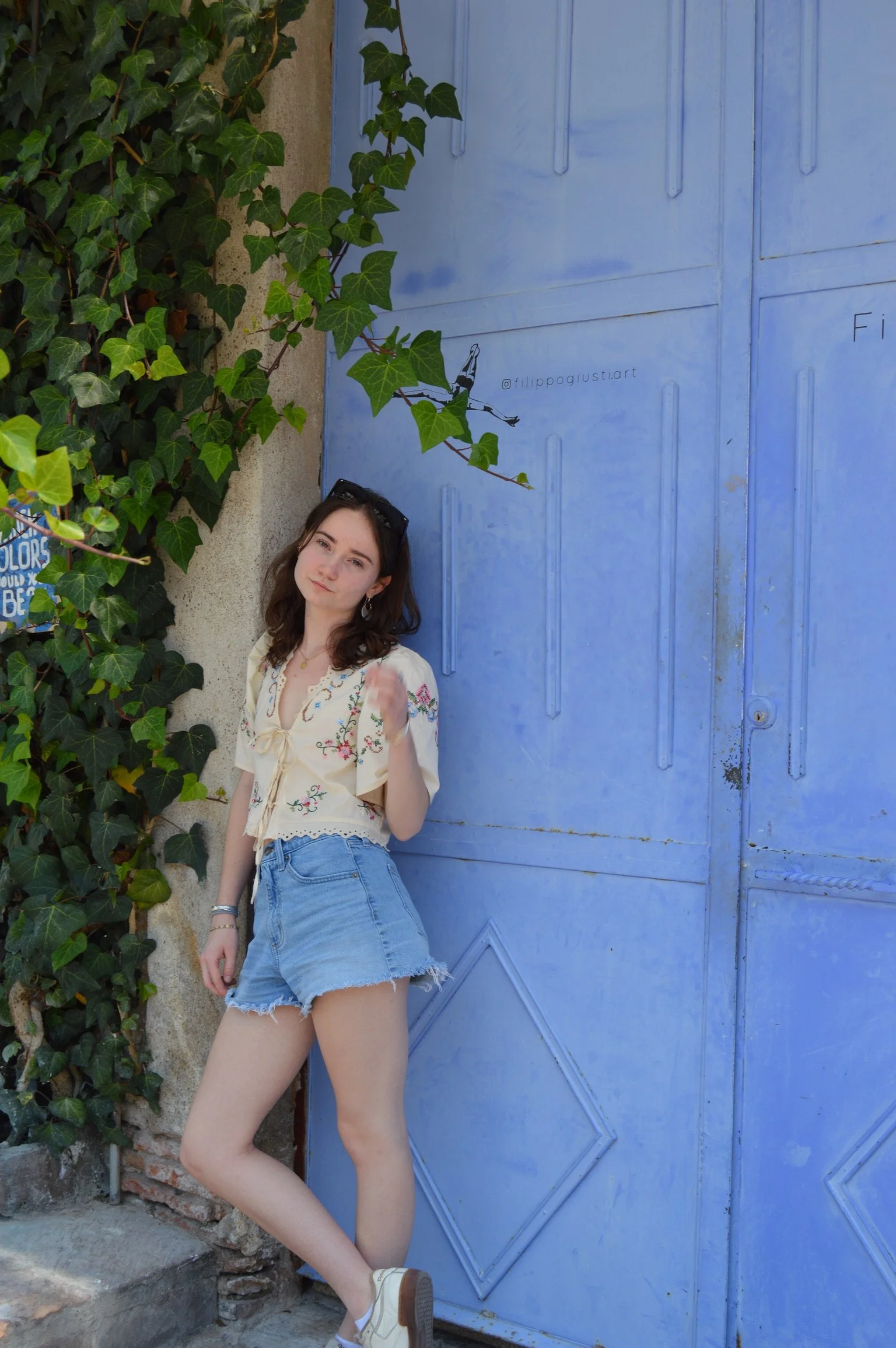 A young woman with shoulder-length brown hair, wearing a cream-colored embroidered blouse, light blue denim shorts, and white sneakers, standing outdoors next to a large blue metal door and a wall covered with green ivy.
