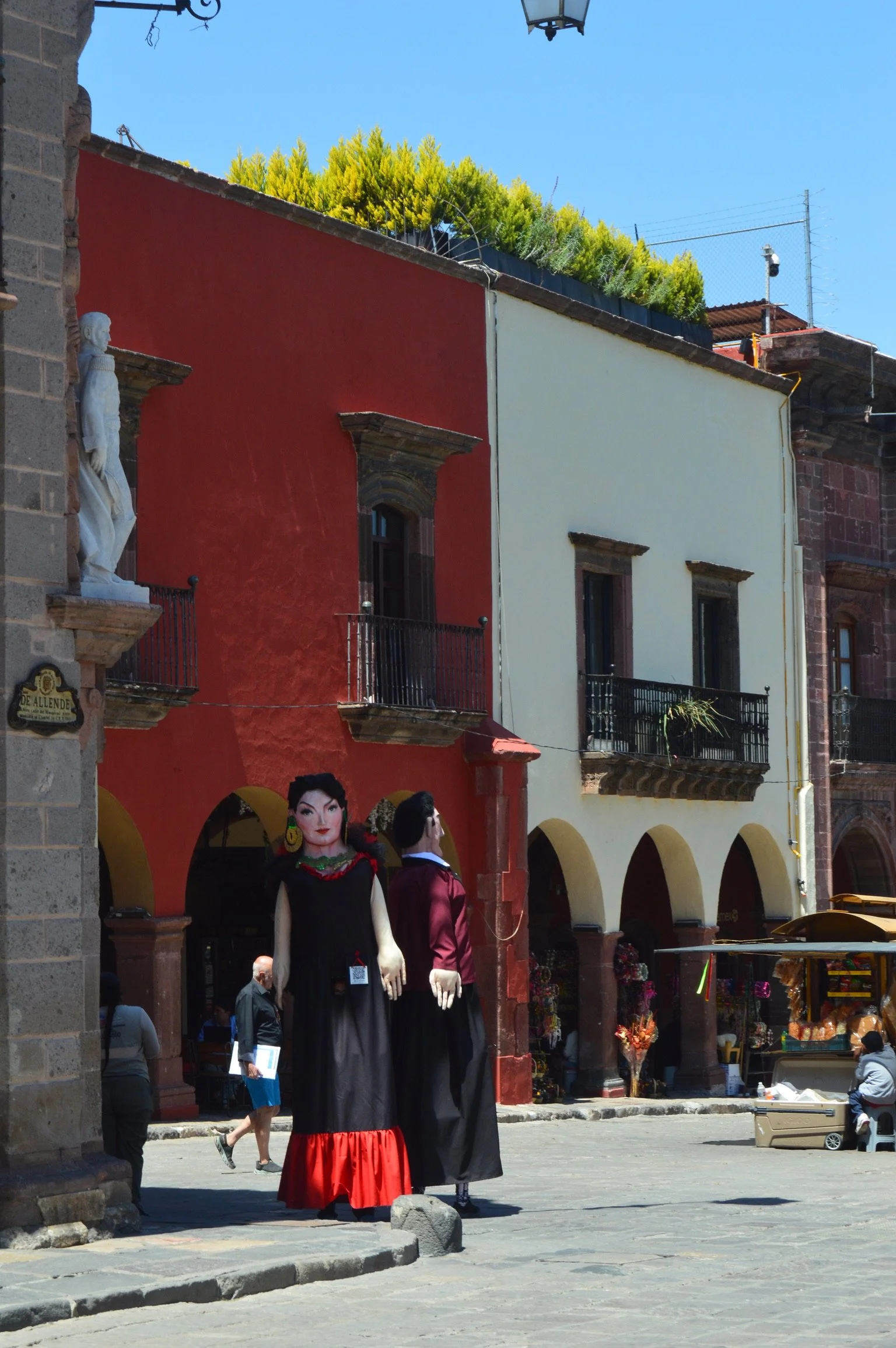 Colorful street scene with traditional buildings, a statue, and giant puppets dressed in folkloric costumes.