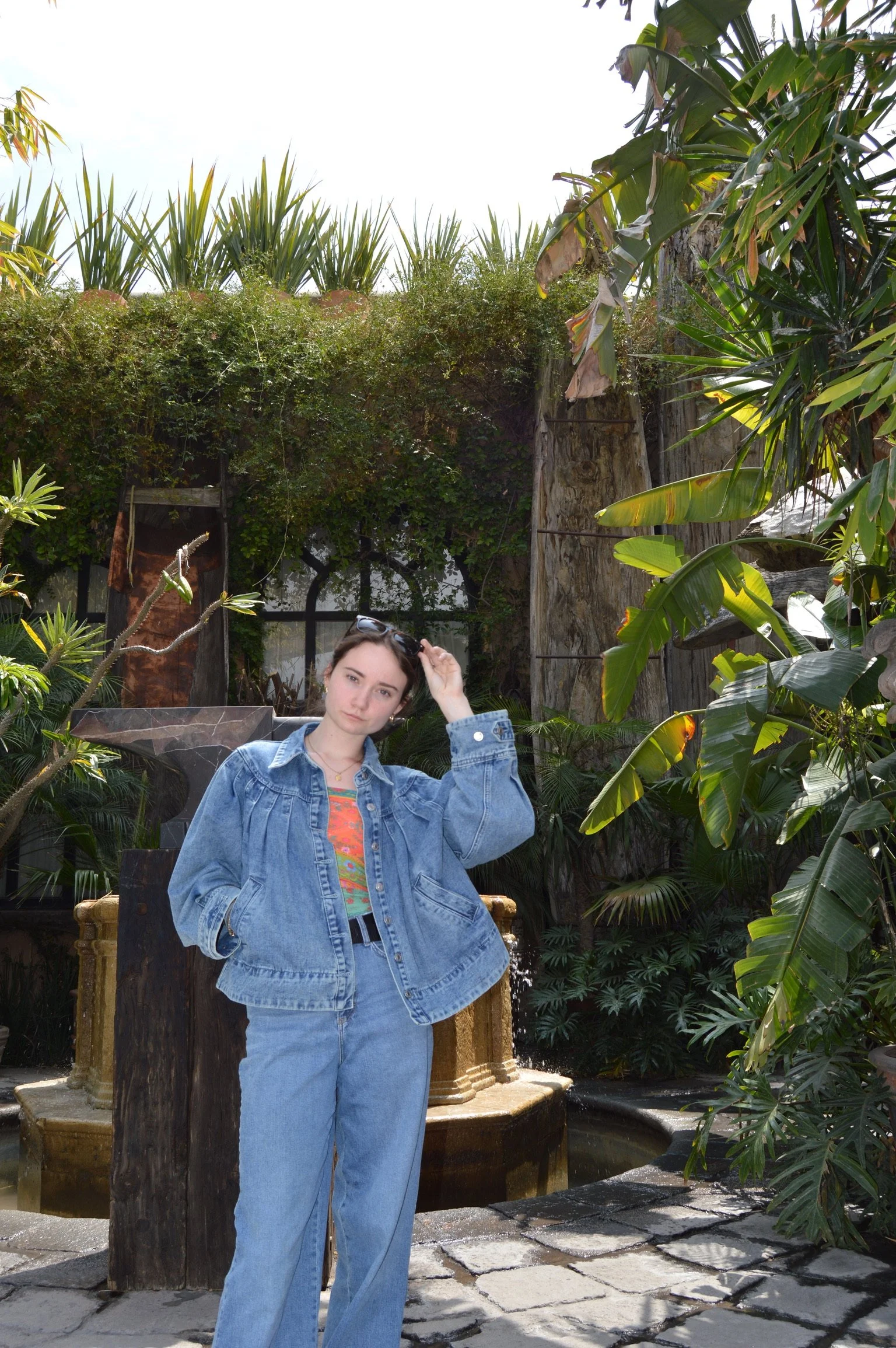 Young woman in denim jacket and jeans standing in lush tropical garden near fountain.